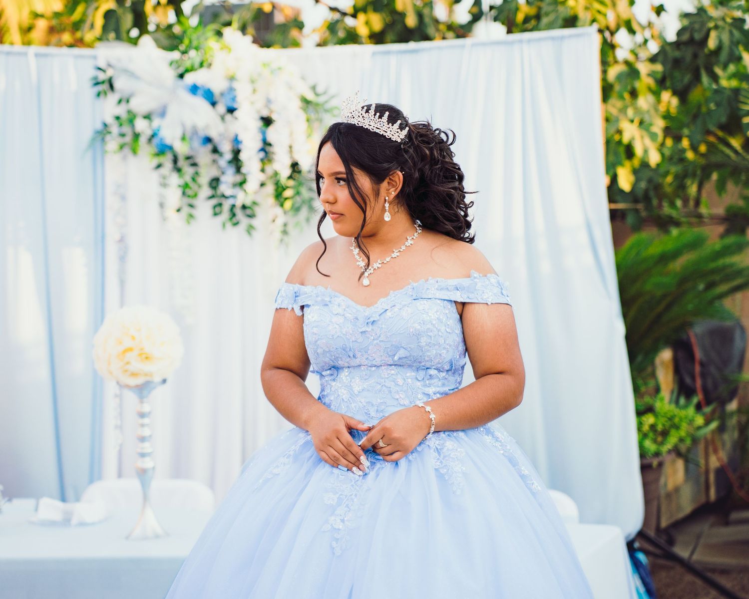 A person in a light blue off-shoulder ballgown poses in front of a white backdrop with floral decorations.