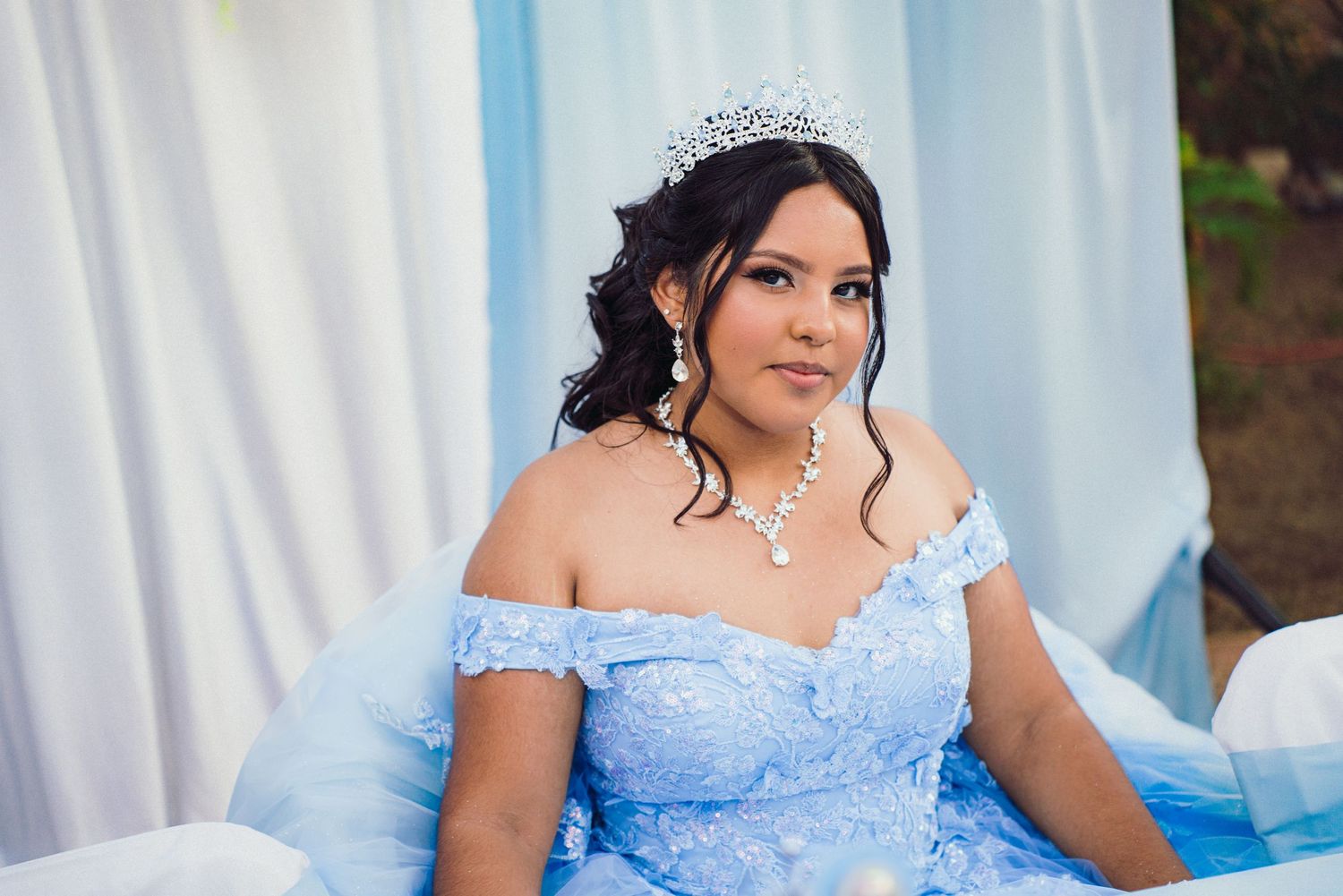 A person wearing a light blue formal dress poses against a backdrop of white curtains and outdoor scenery.