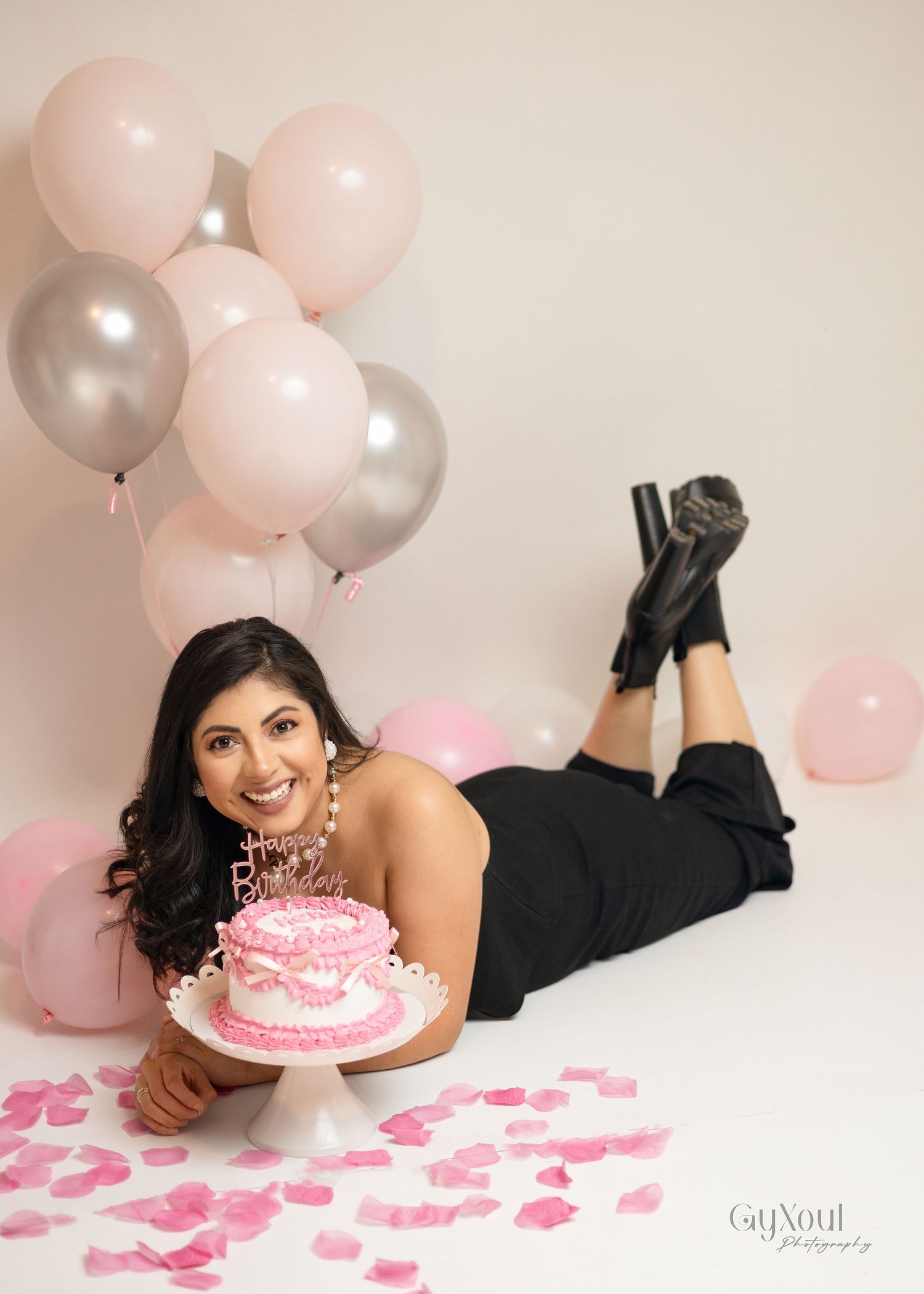 Pink and silver balloons create a romantic backdrop for a birthday celebration with cake and rose petals on the floor.