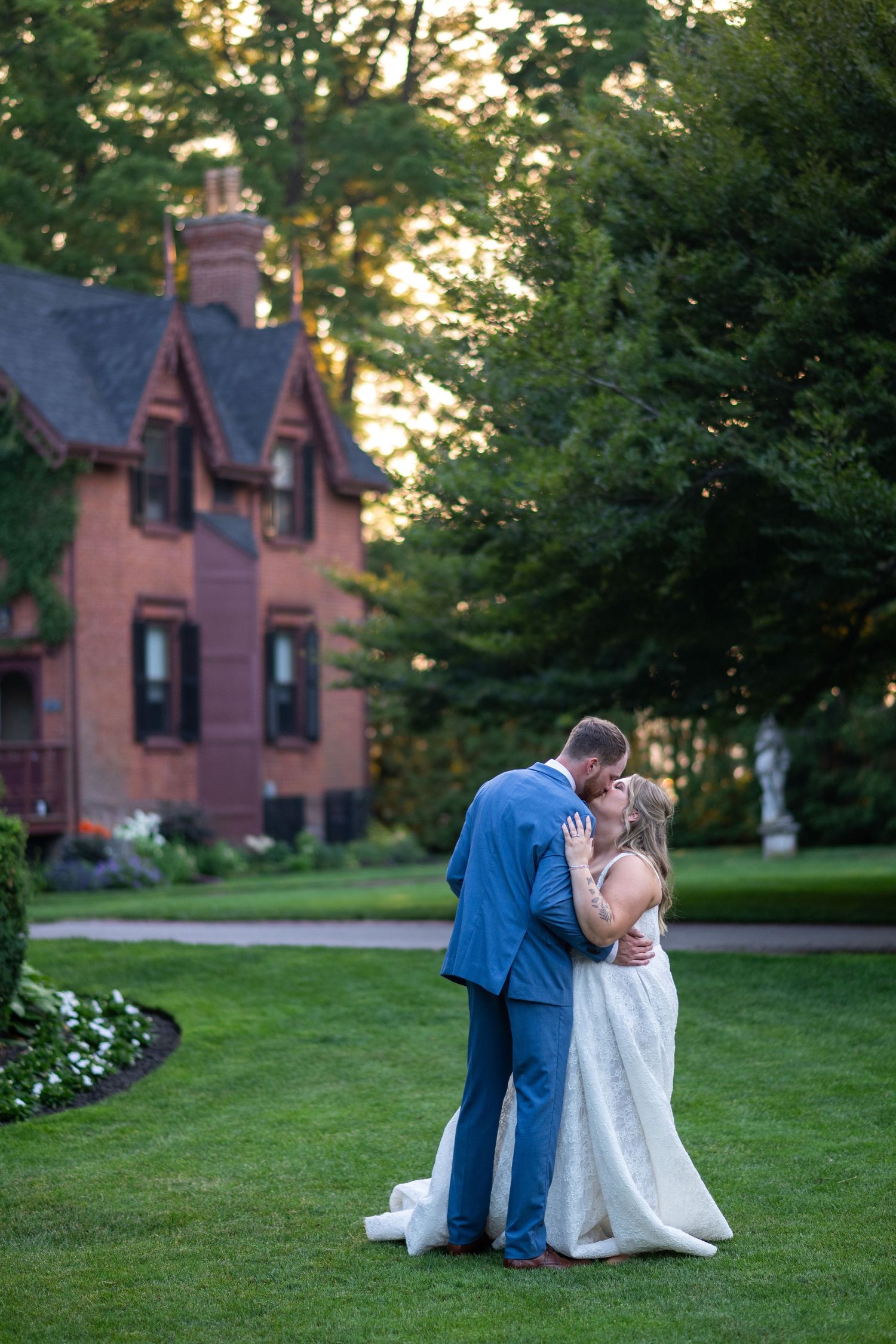 A romantic moment unfolds on a lush green lawn with a historic brick mansion in the background at sunset.