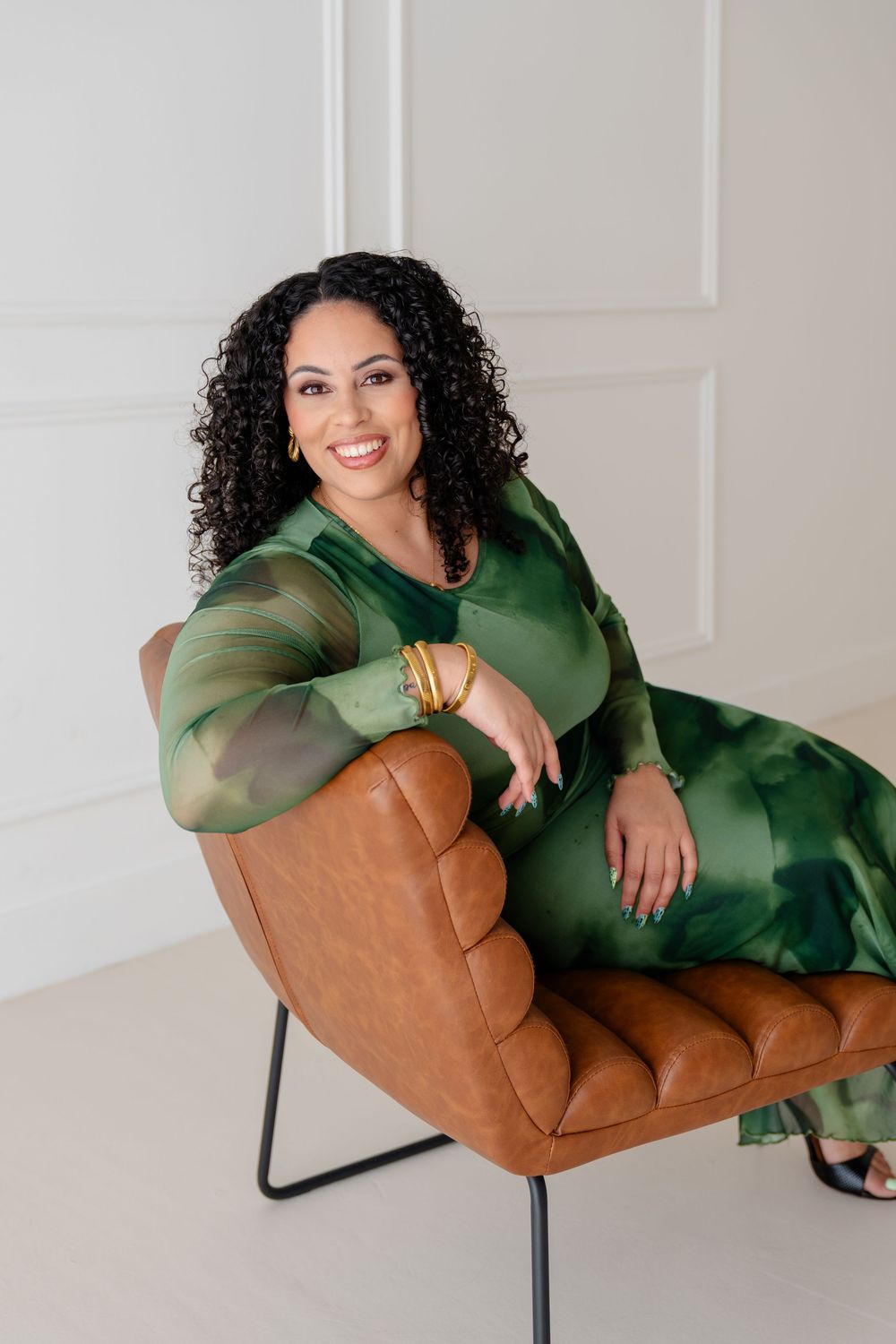 A person in a green dress lounges on a modern brown leather chair against a white wall.