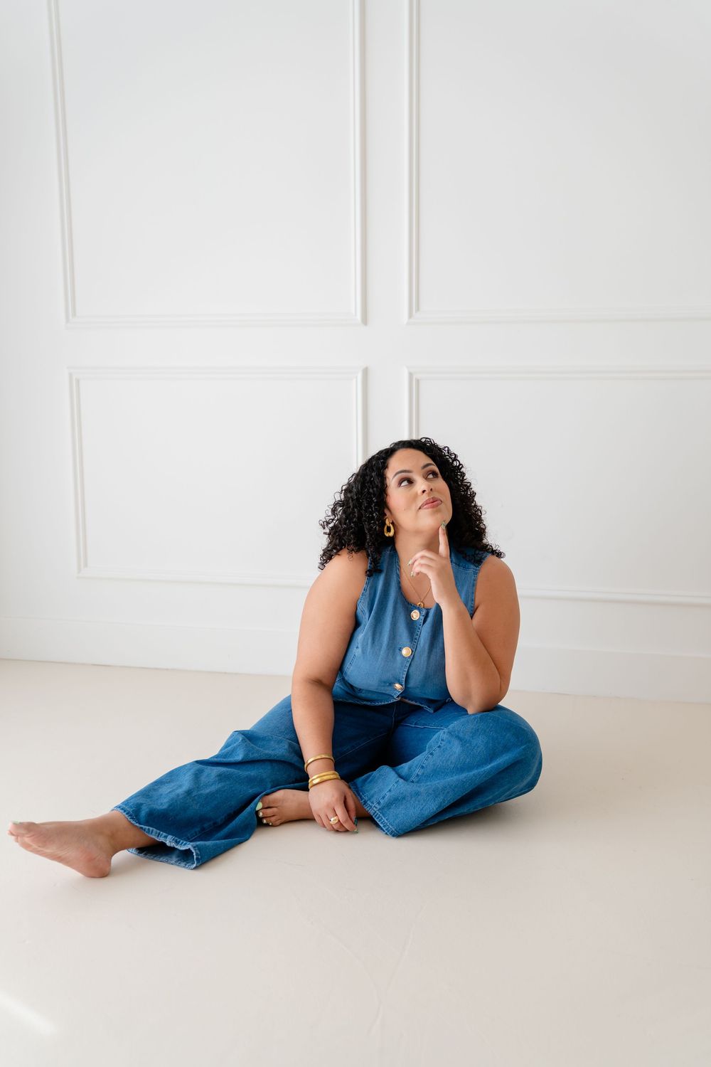 Someone in a blue denim outfit sits cross-legged on a white floor against a white wall, looking upward thoughtfully.