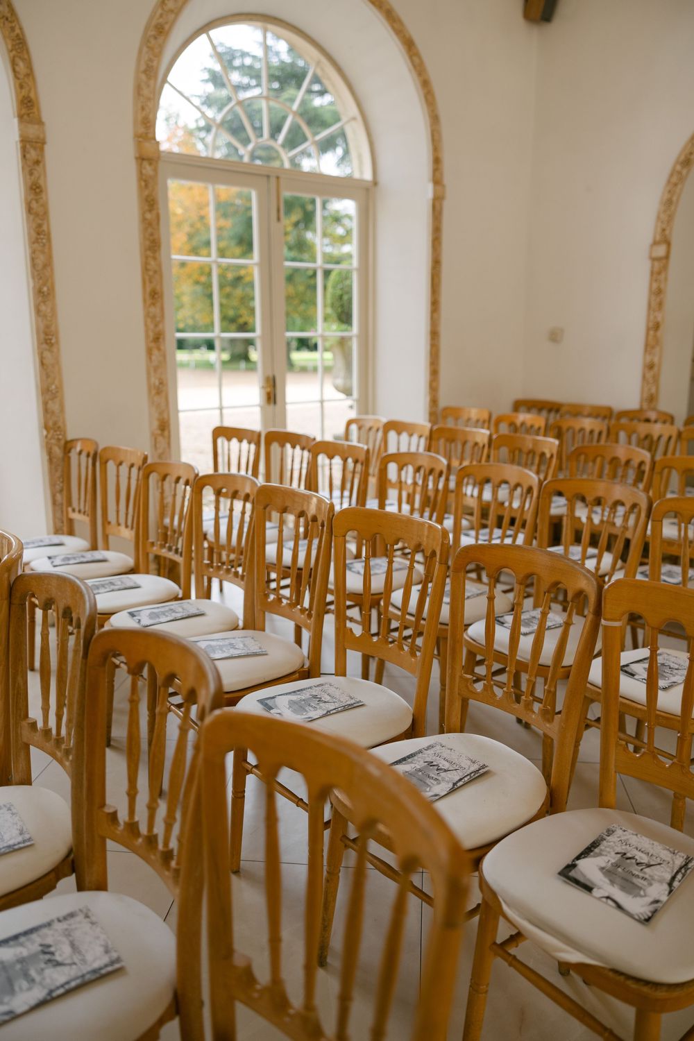 Elegant wedding venue setup with rows of golden chairs and white cushions in a bright room with arched windows.