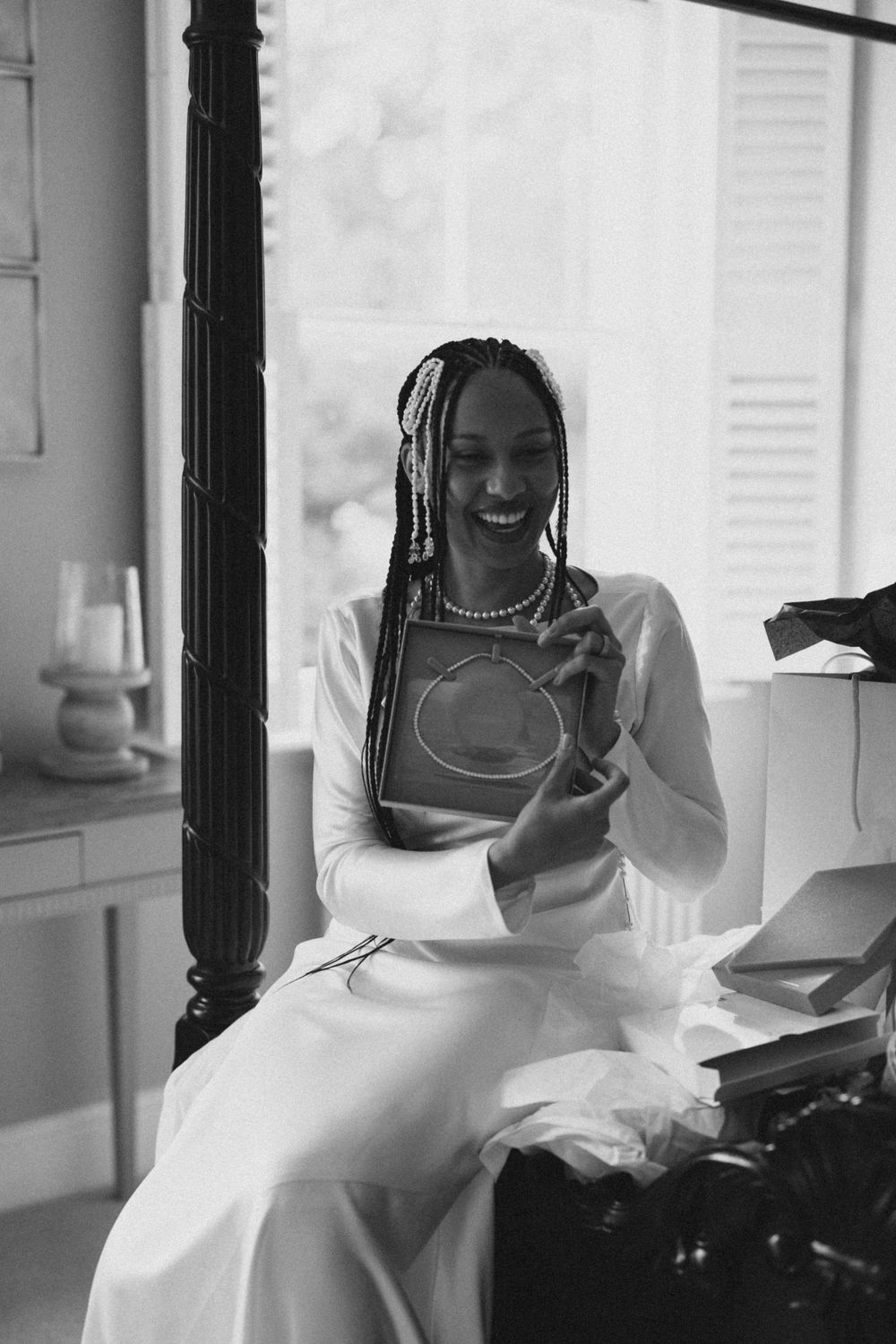 A person in a white outfit with braided hair sits smiling while holding a book in a black and white photograph.