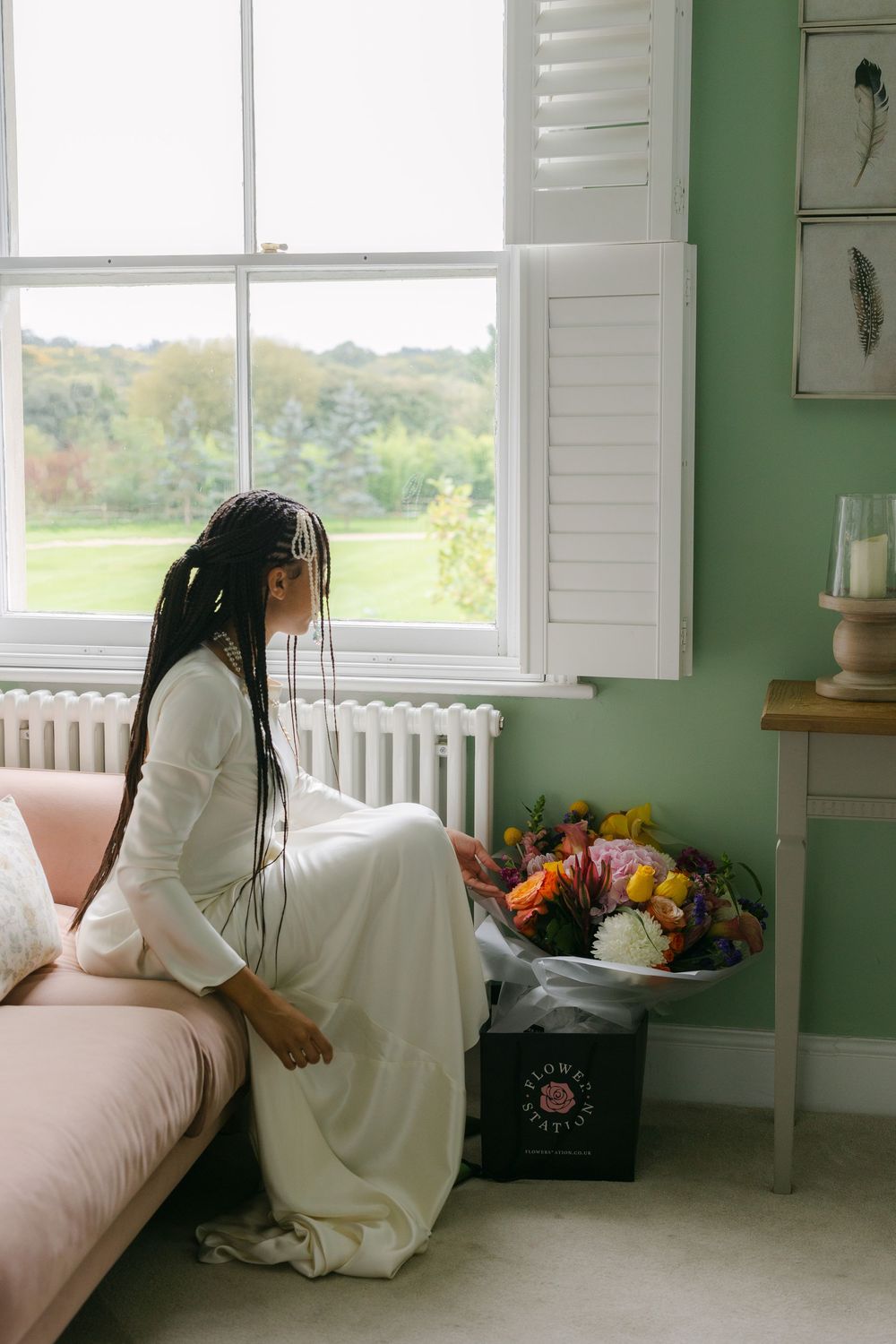 A figure in a flowing white dress sits by a window in a mint green room with natural light streaming in.