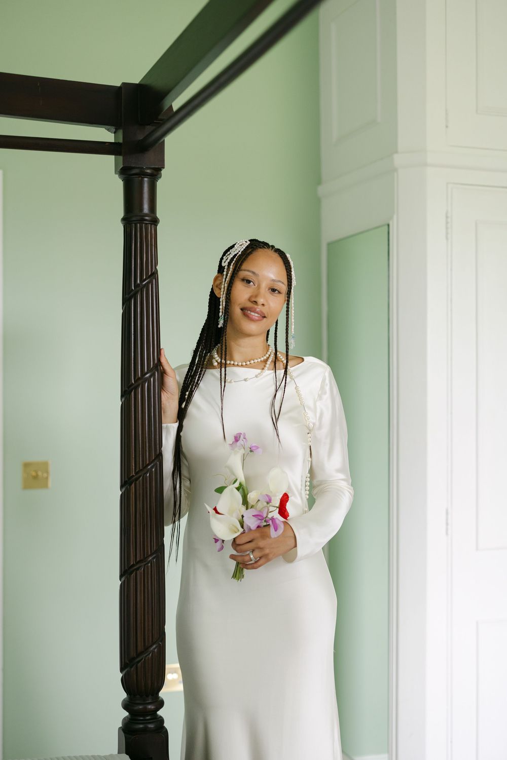 A person in a white long-sleeved dress poses against a mint green wall next to a dark wooden pillar.