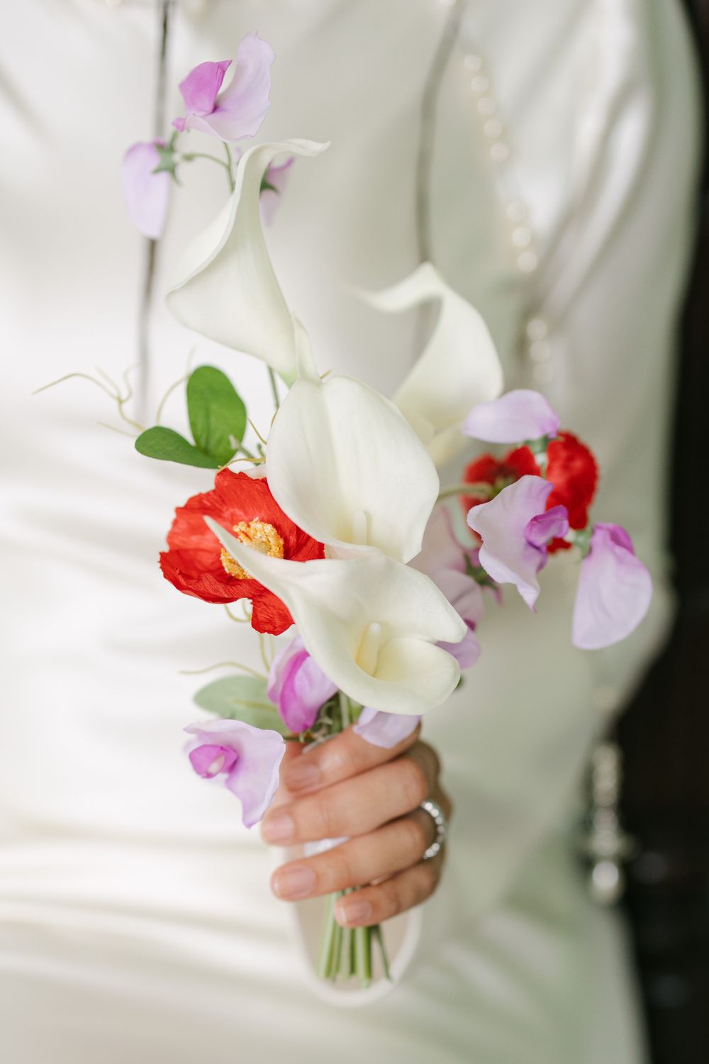 A delicate bridal bouquet featuring white calla lilies, pink sweet peas and red poppies held against a white dress.