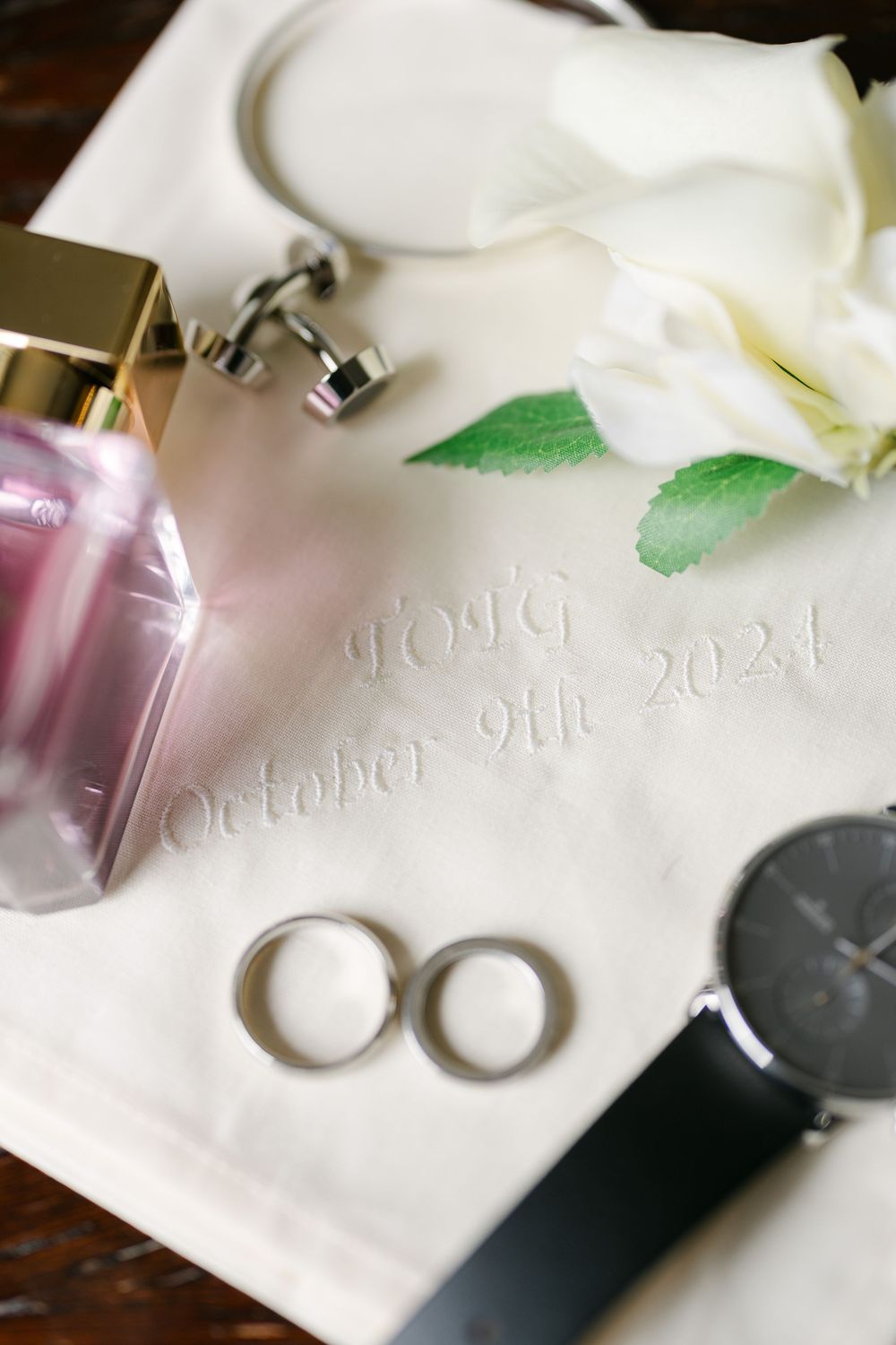 Wedding details including two silver rings, a perfume bottle, watch, and white calla lily on embossed paper with a date.