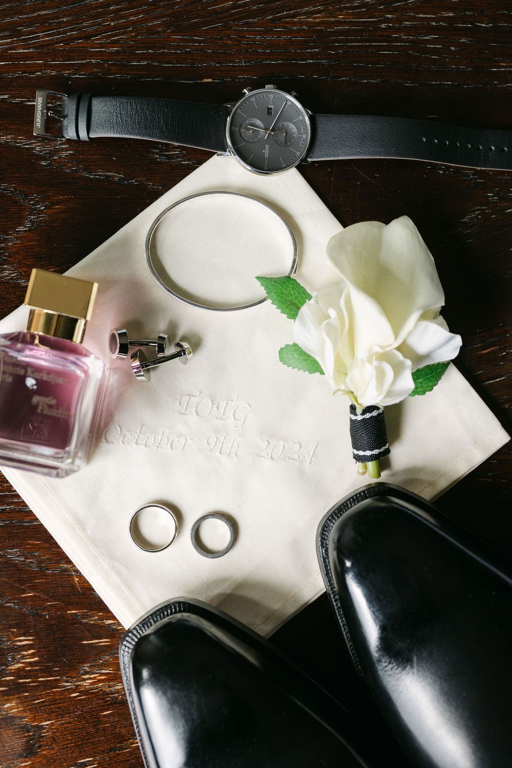 Black leather dress shoes with white boutonniere flowers and greenery on a dark wooden surface.