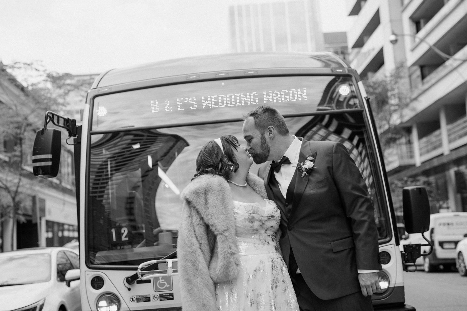 Newlyweds sharing a kiss in front of their wedding wagon bus on a city street in black and white.