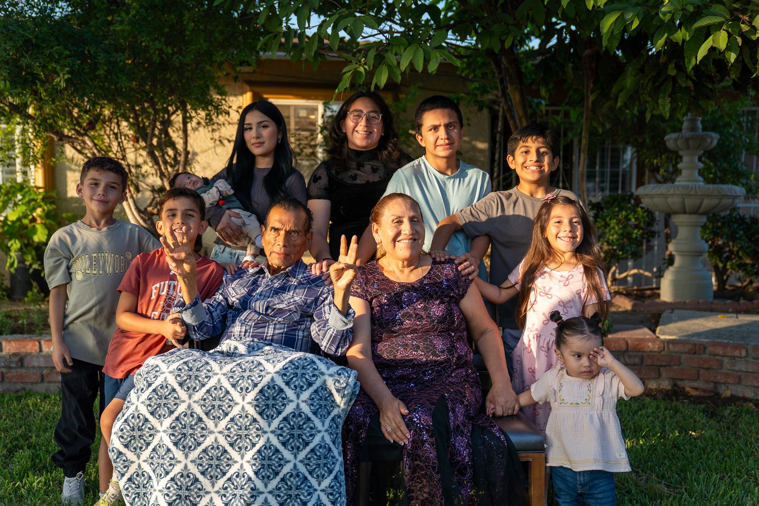 A family gathering outdoors with green foliage and a blue patterned pillow visible in the background.