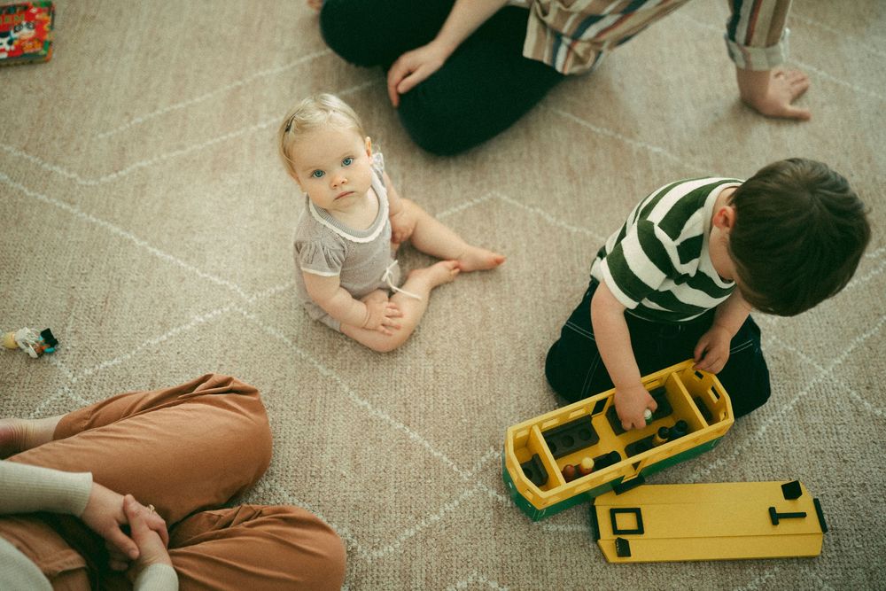 Children playing on the floor with a yellow toy construction vehicle while adults sit nearby.