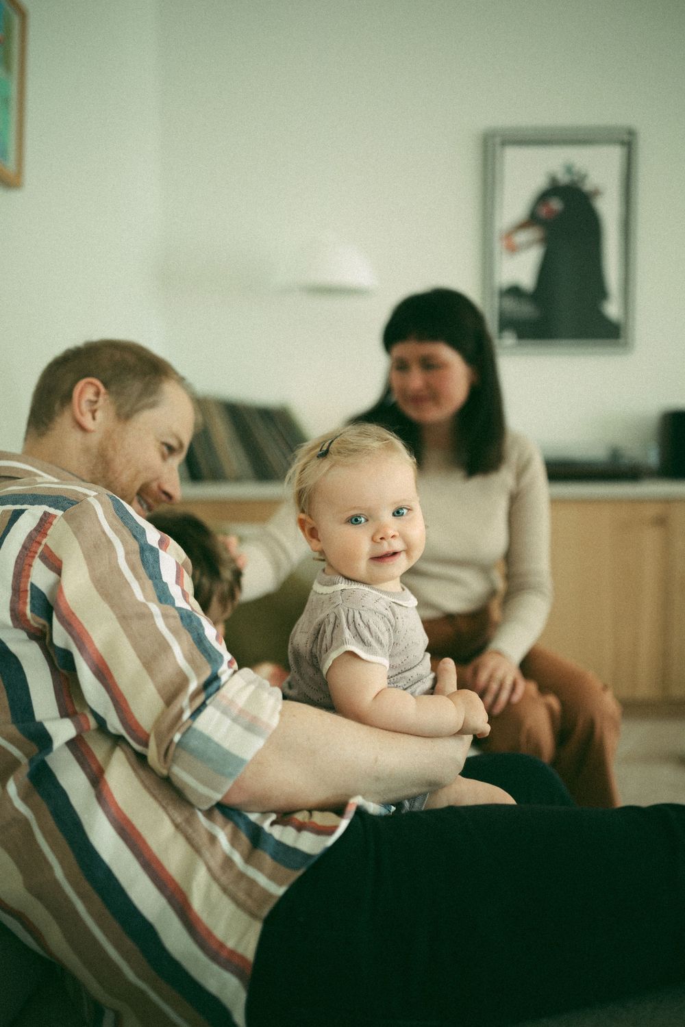 A baby in a striped shirt plays on a couch while an adult sits in the background of a living room.