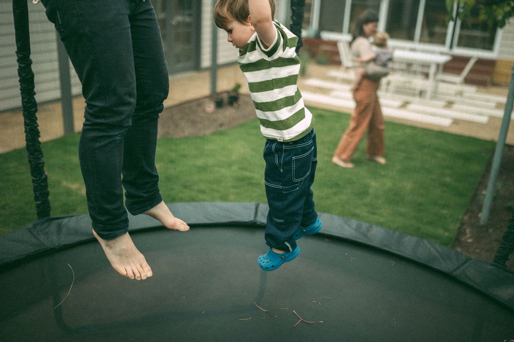 Bare feet and child's feet jumping on backyard trampoline with green grass and patio furniture visible in background.