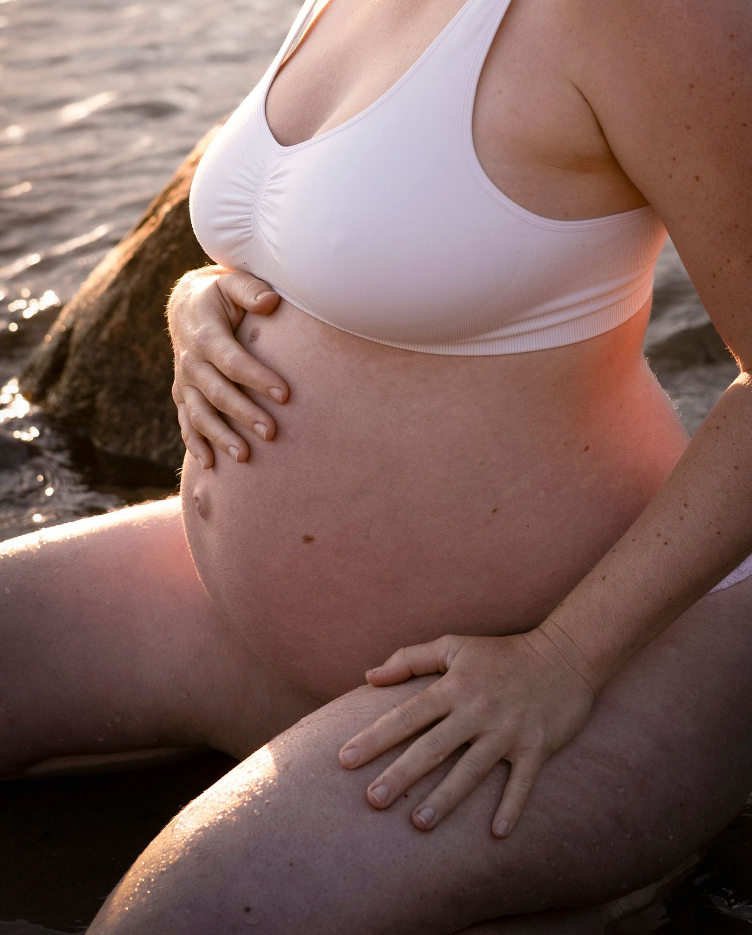 gravid kvinna sitter på knä vid havskanten. hon håller sig om magen. hon är klädd i vitt.