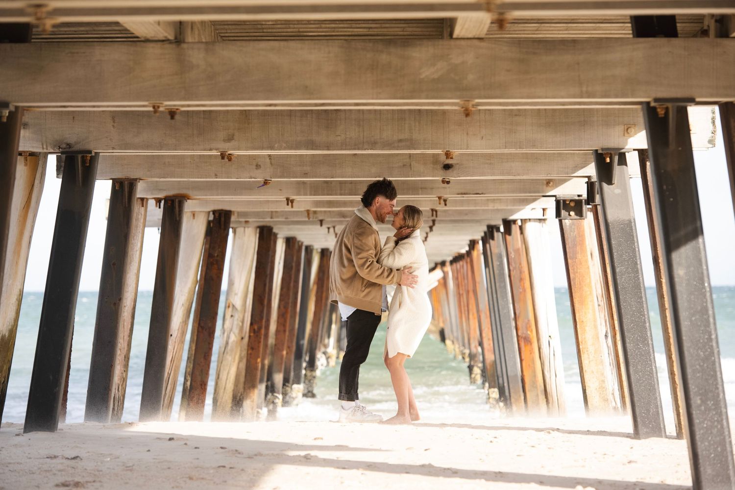 Couple sharing a romantic embrace under a weathered beach pier with sandy ground and sunlight filtering through the columns.