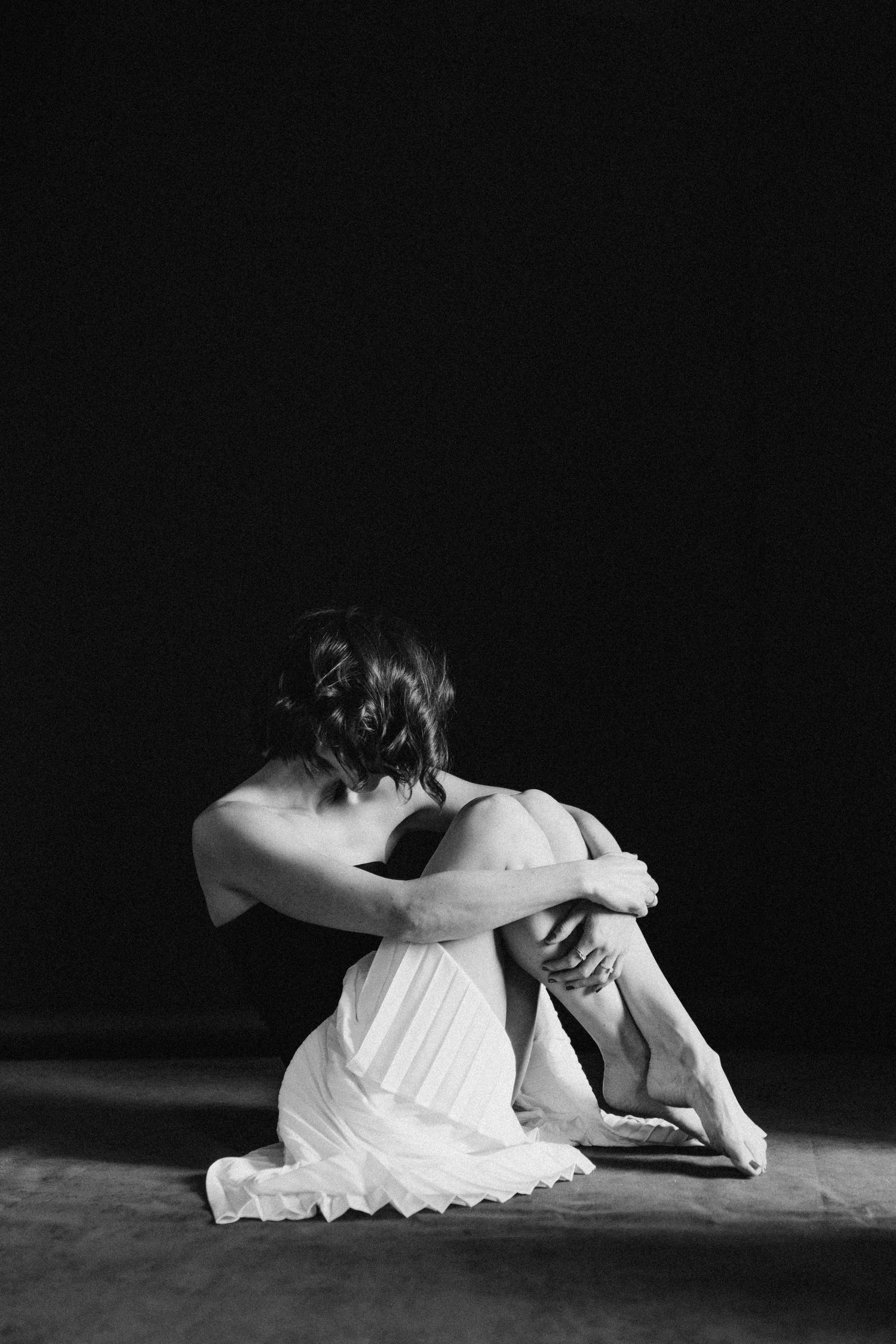 Black and white photo of a woman in a white dress sitting on the floor hugging her knees in a dark studio.