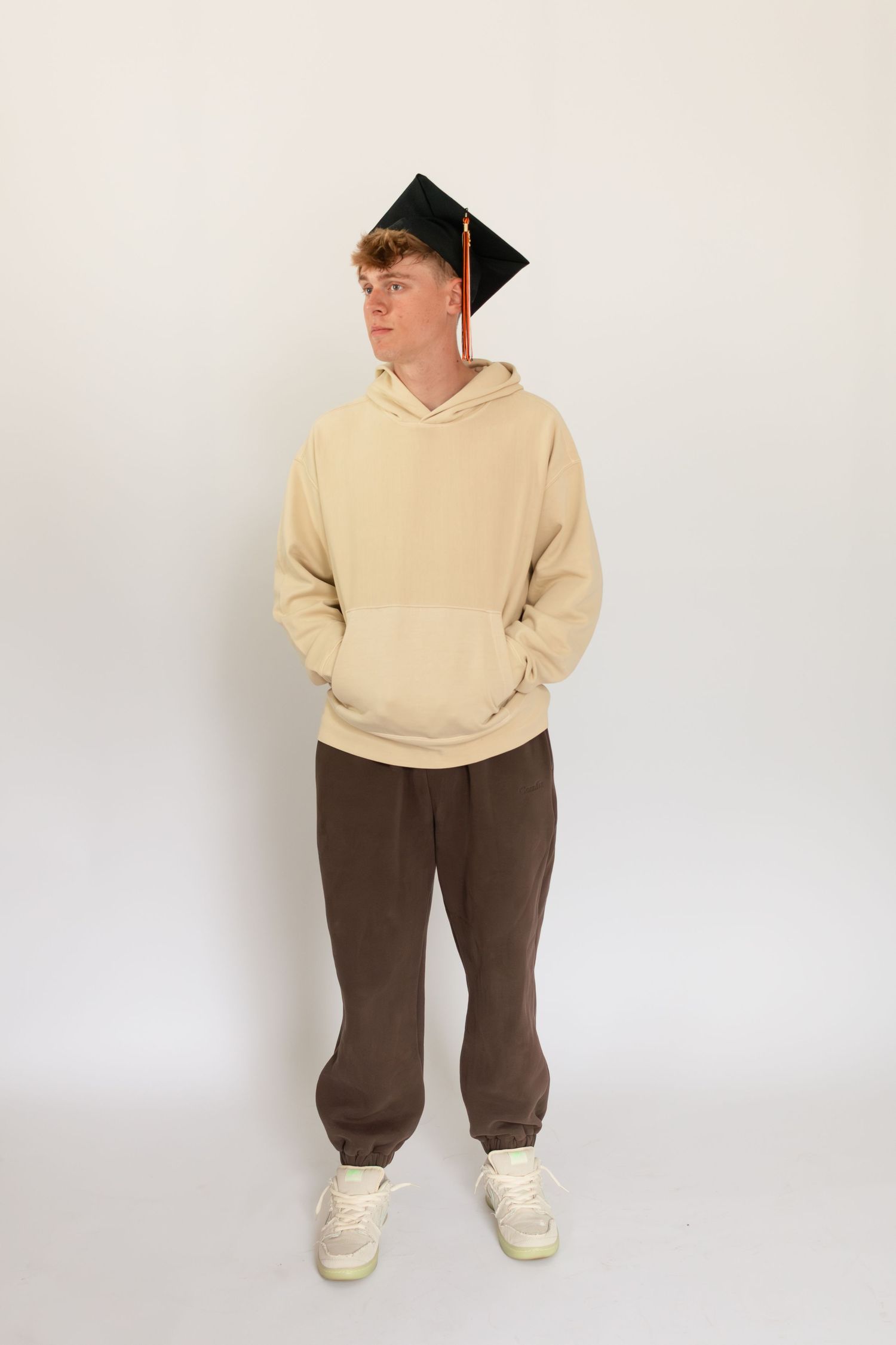 Graduate wearing a beige hoodie and brown sweatpants stands against white background with graduation cap.Family, senior, and wedding photogr