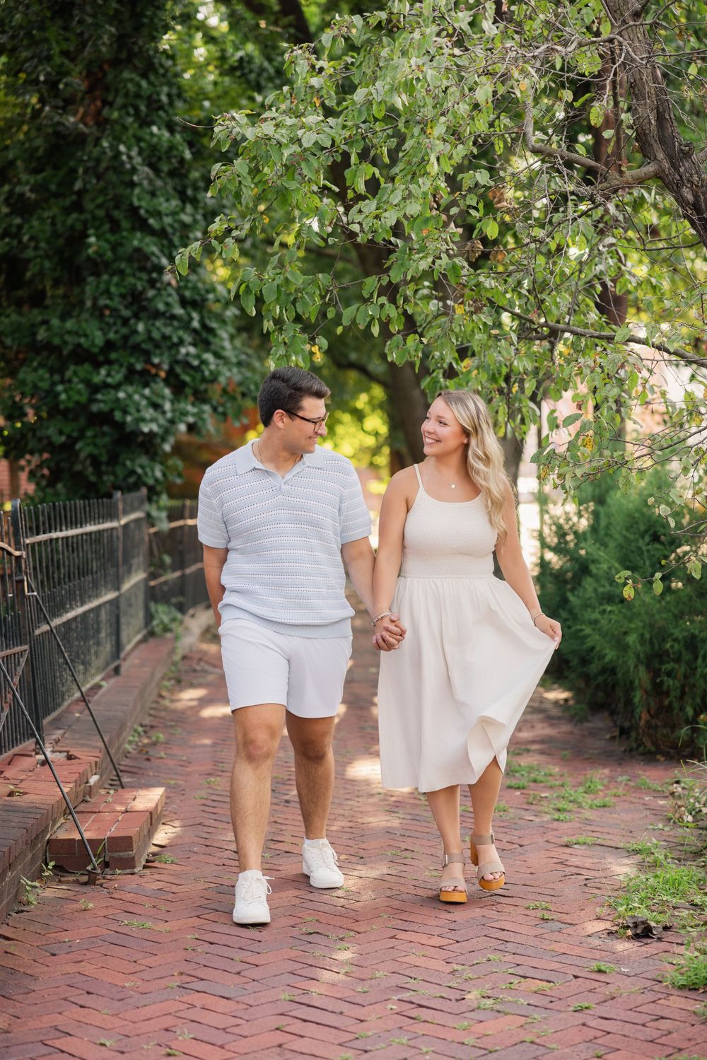 A couple walking hand in hand along a brick pathway surrounded by lush greenery on a sunny day. German Village Columbus Ohio Engagement