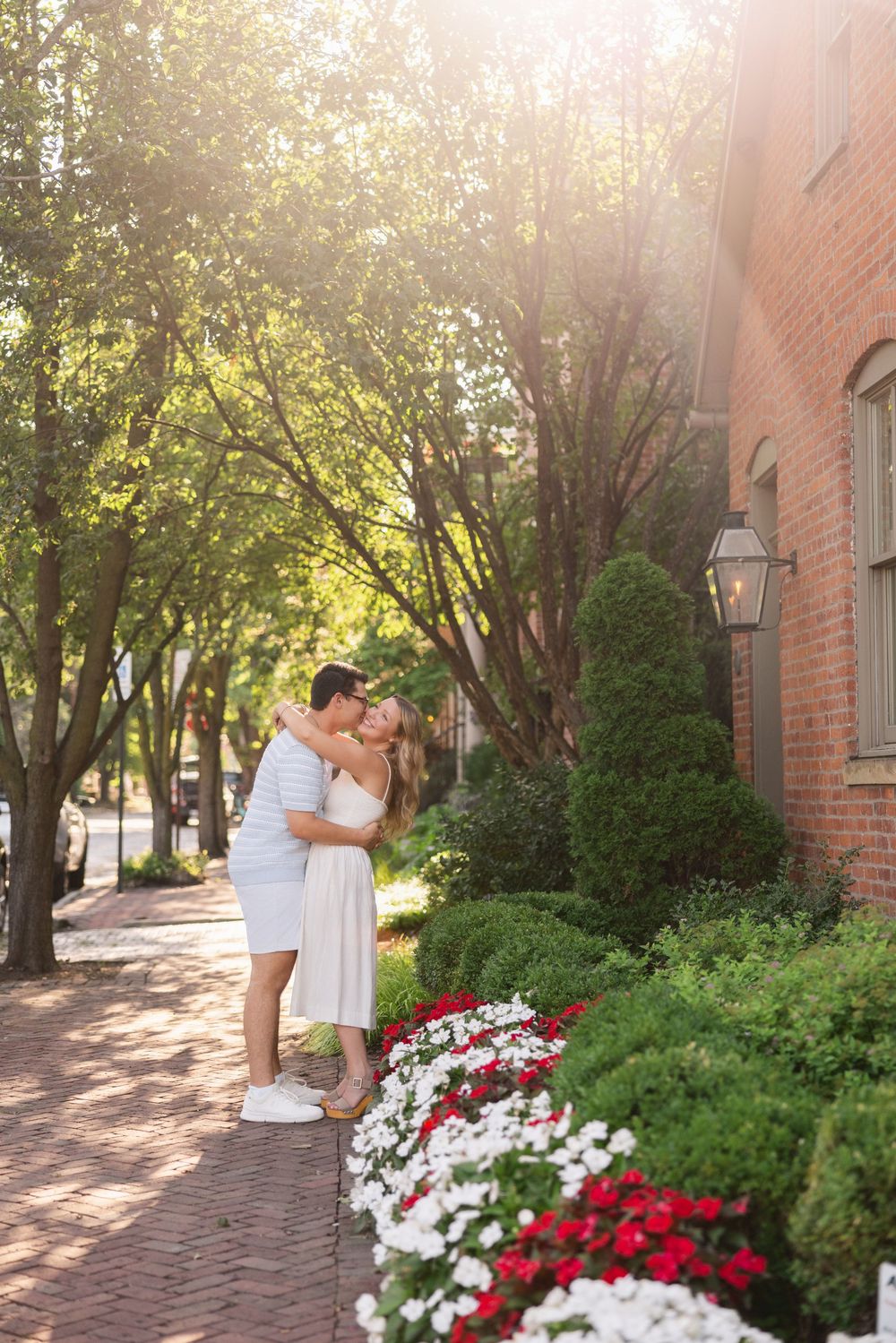 A romantic couple kisses near red and white flowers along a brick building with beautiful sunlit trees. German Village Columbus Ohio