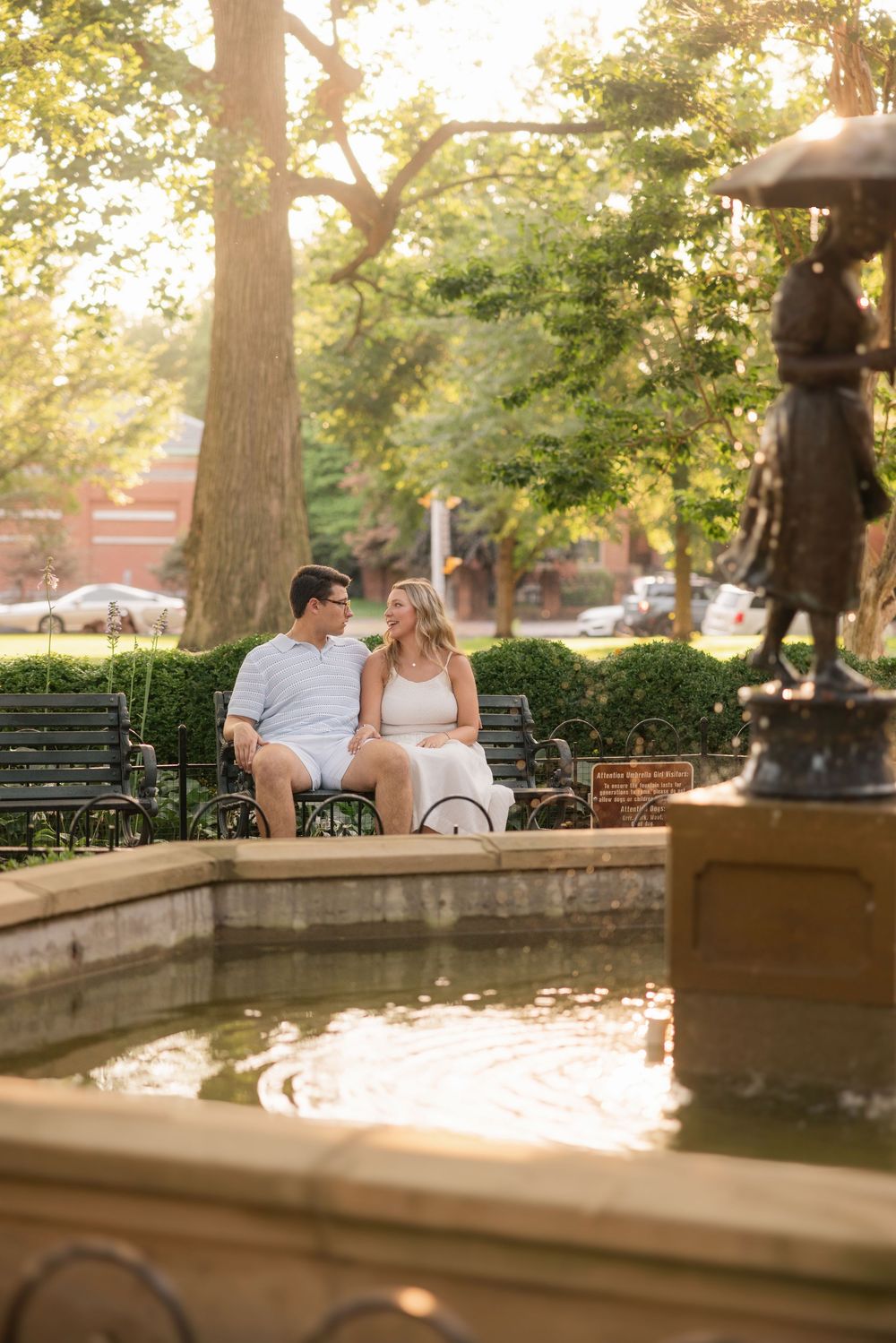 A couple sits together on a park bench near a fountain during golden hour. Schiller Park Engagement Session Columbus Ohio.