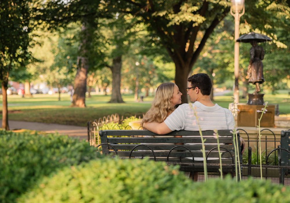 A couple sitting together on a park bench surrounded by trees in warm afternoon sunlight. German Village Columbus Ohio Engagement Session