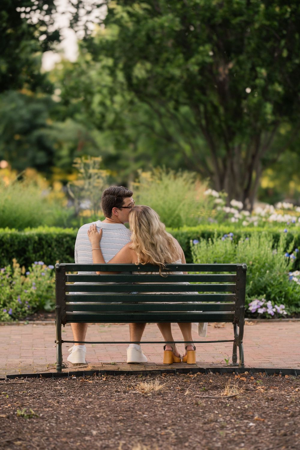 A couple shares an intimate moment while sitting on a park bench surrounded by lush gardens. Schiller Park Engagement Photo.