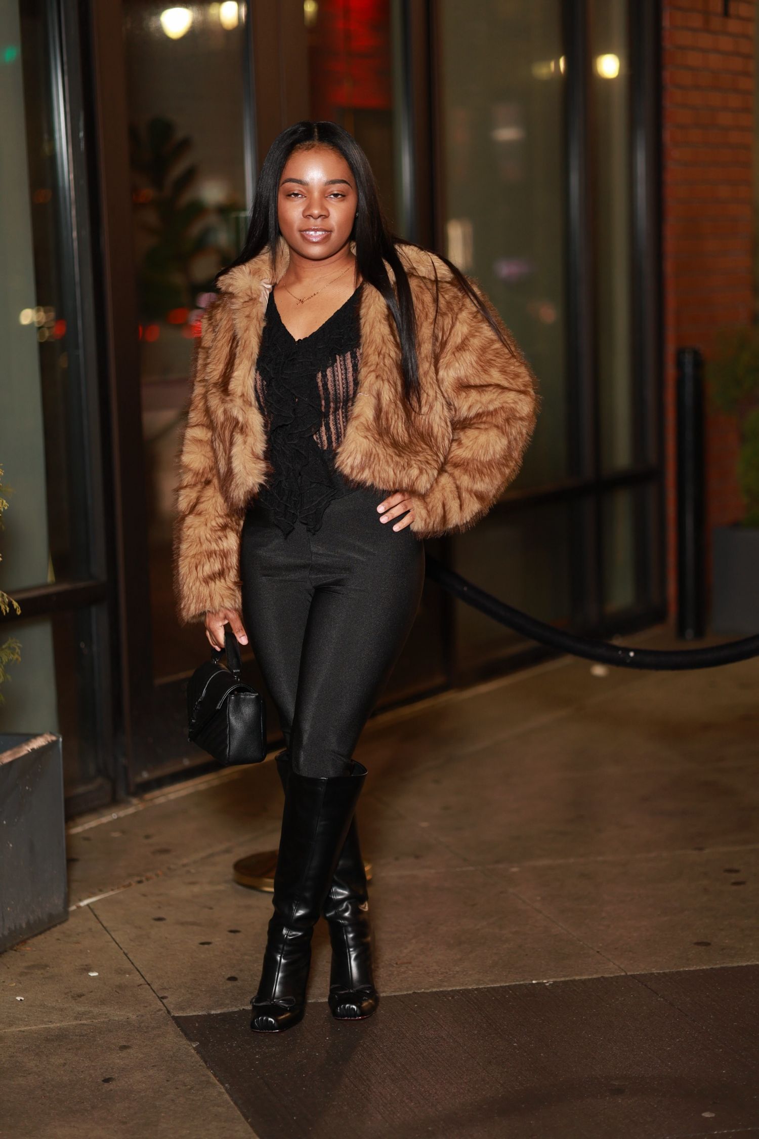 A woman walks at night wearing a brown fur coat, black pants and knee-high boots with a small handbag.