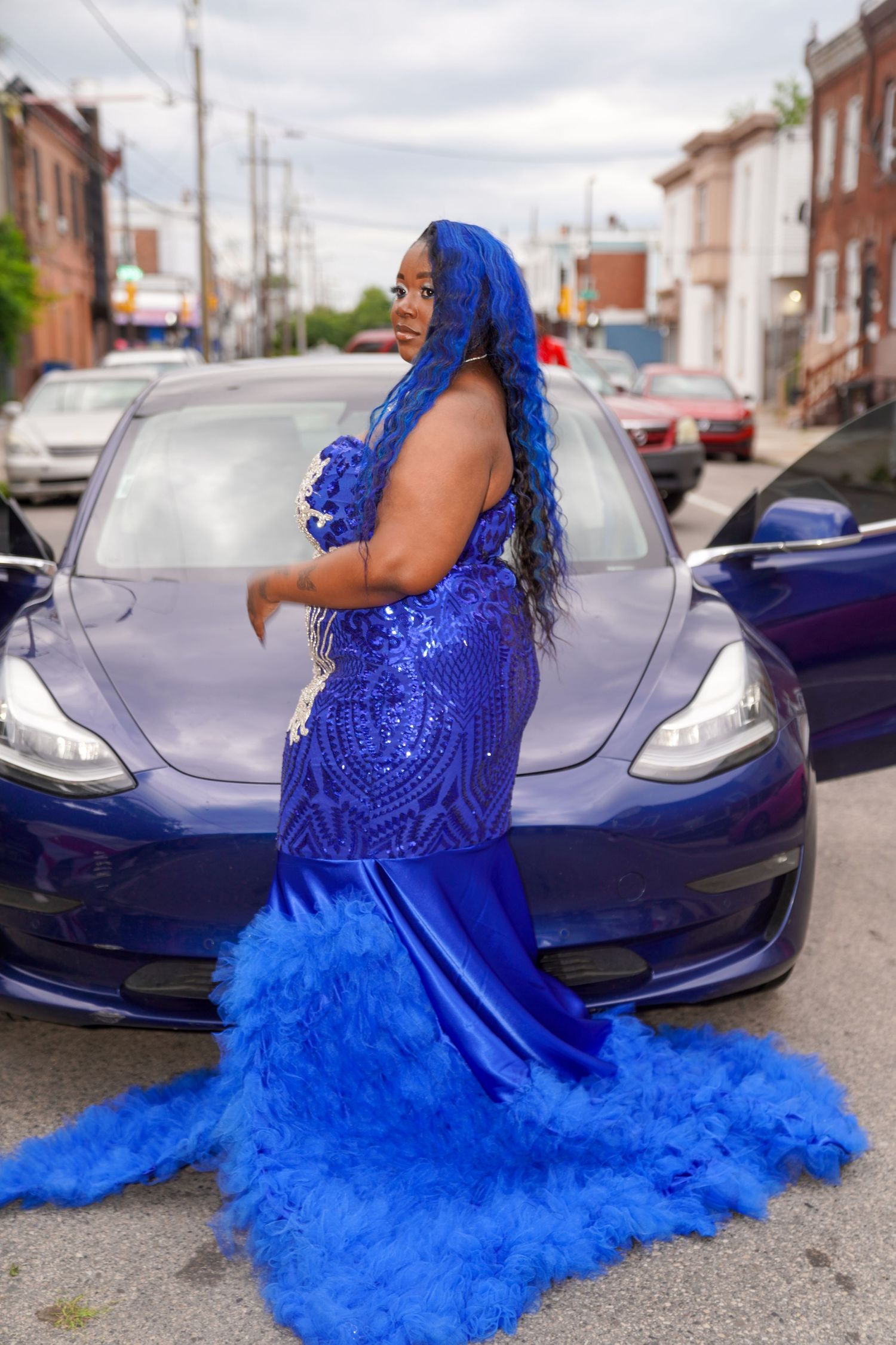 A vibrant royal blue tulle gown with lace details photographed against a Tesla Model 3 car.
