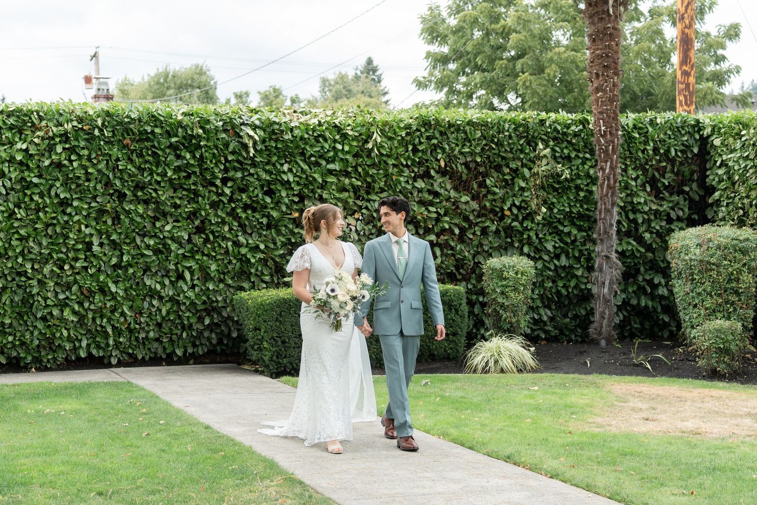 A couple in wedding attire walks through a lush garden path with tall hedges and trees.
