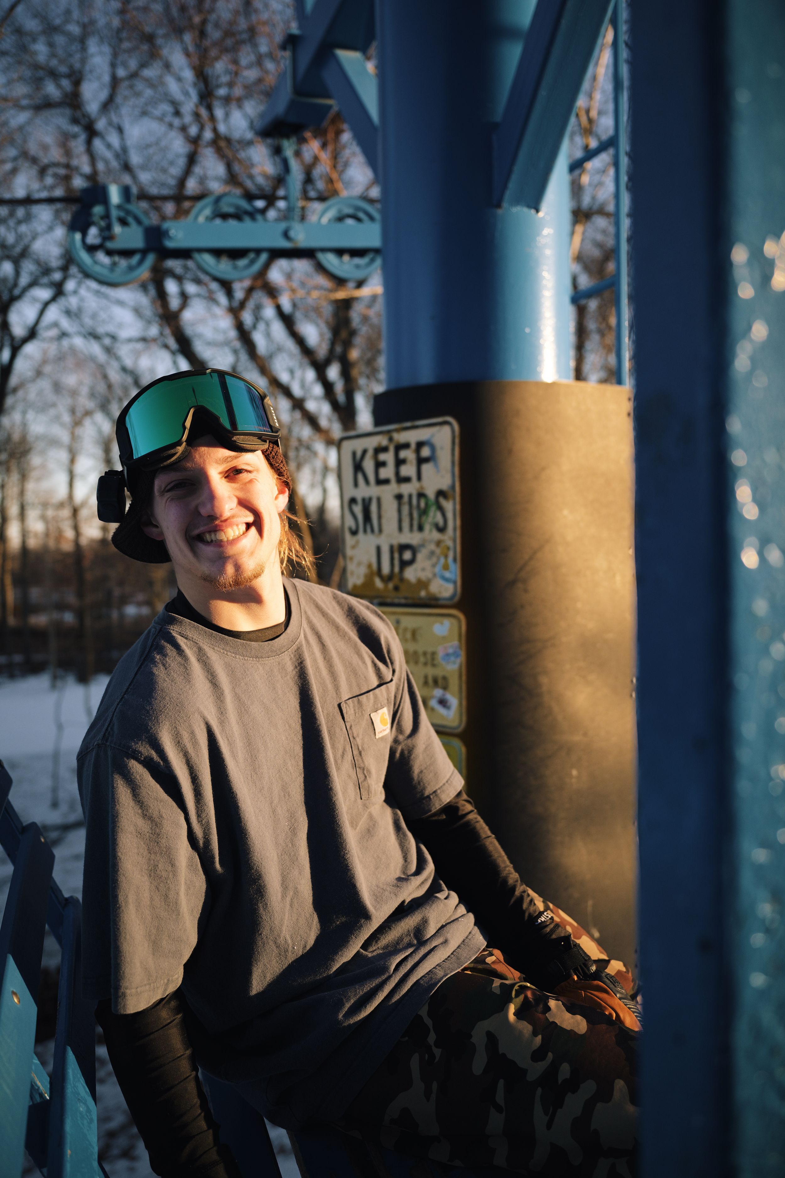 Smiling snowboarder in green goggles leans against blue ski lift structure near Keep Ski Tips Up sign.