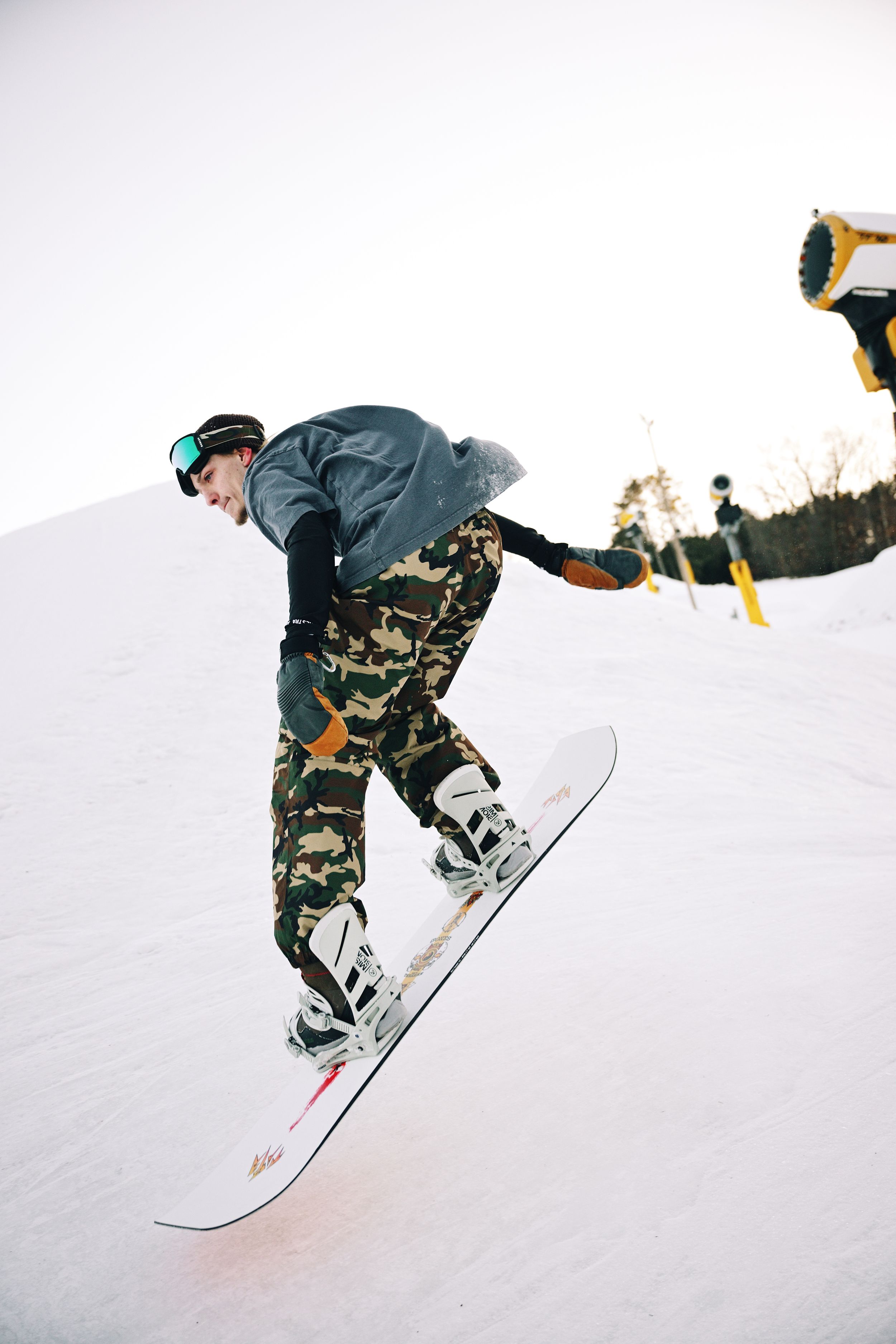 Snowboarder in camouflage pants and goggles performing a trick on a snowy ski slope.