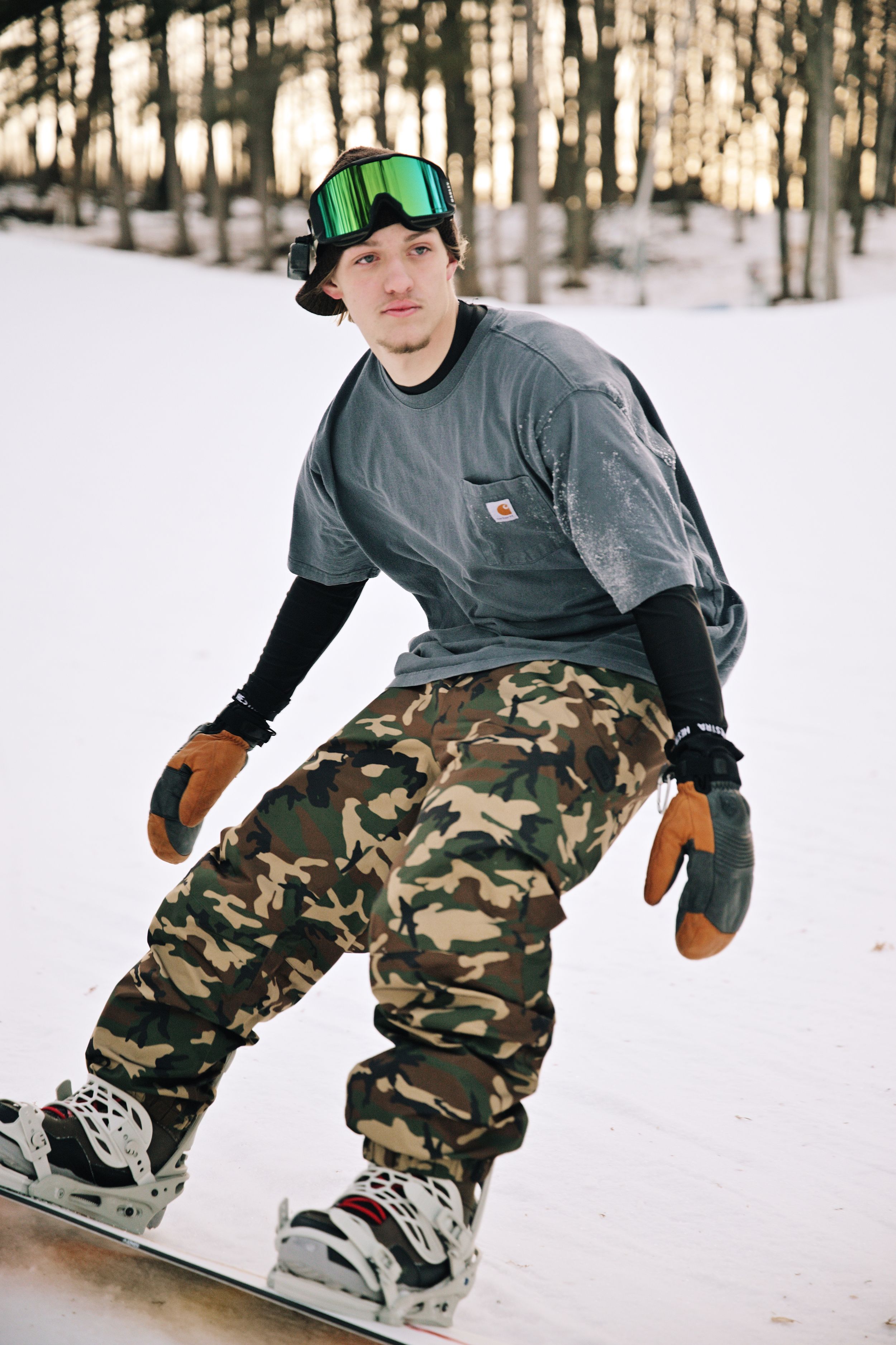 Snowboarder in Carhartt shirt, camo pants, and green goggles rides down a snowy wooded slope.