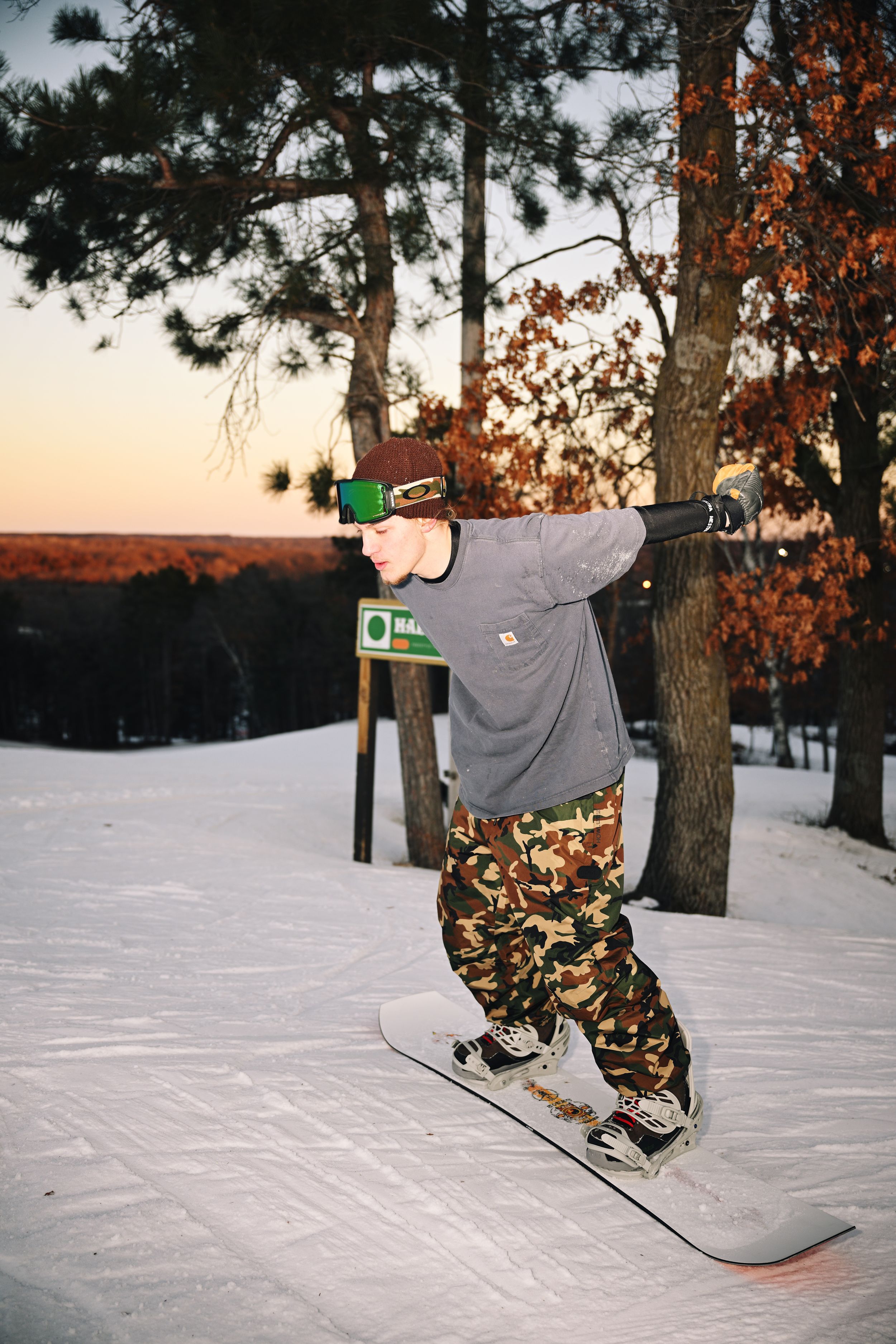 Snowboarder in camo pants balances on board near pine trees on a snowy slope at sunset.