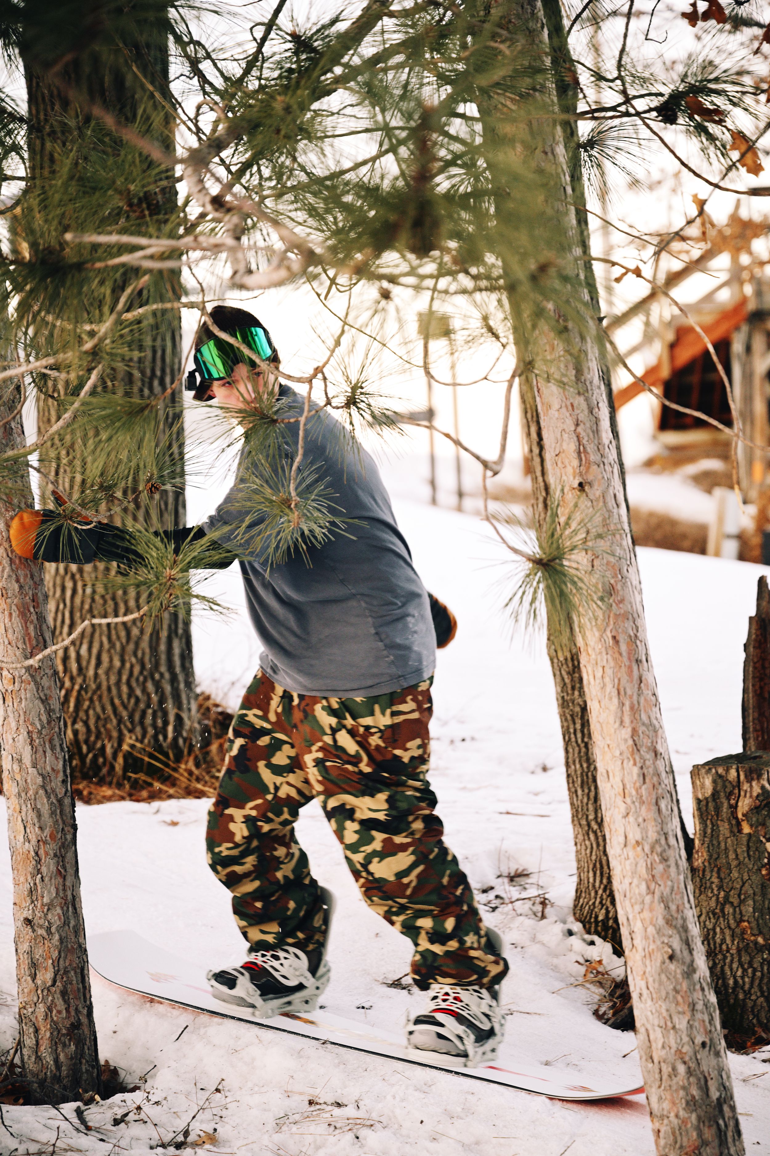 Snowboarder in camo pants navigates through pine trees on a snowy ski slope in winter.