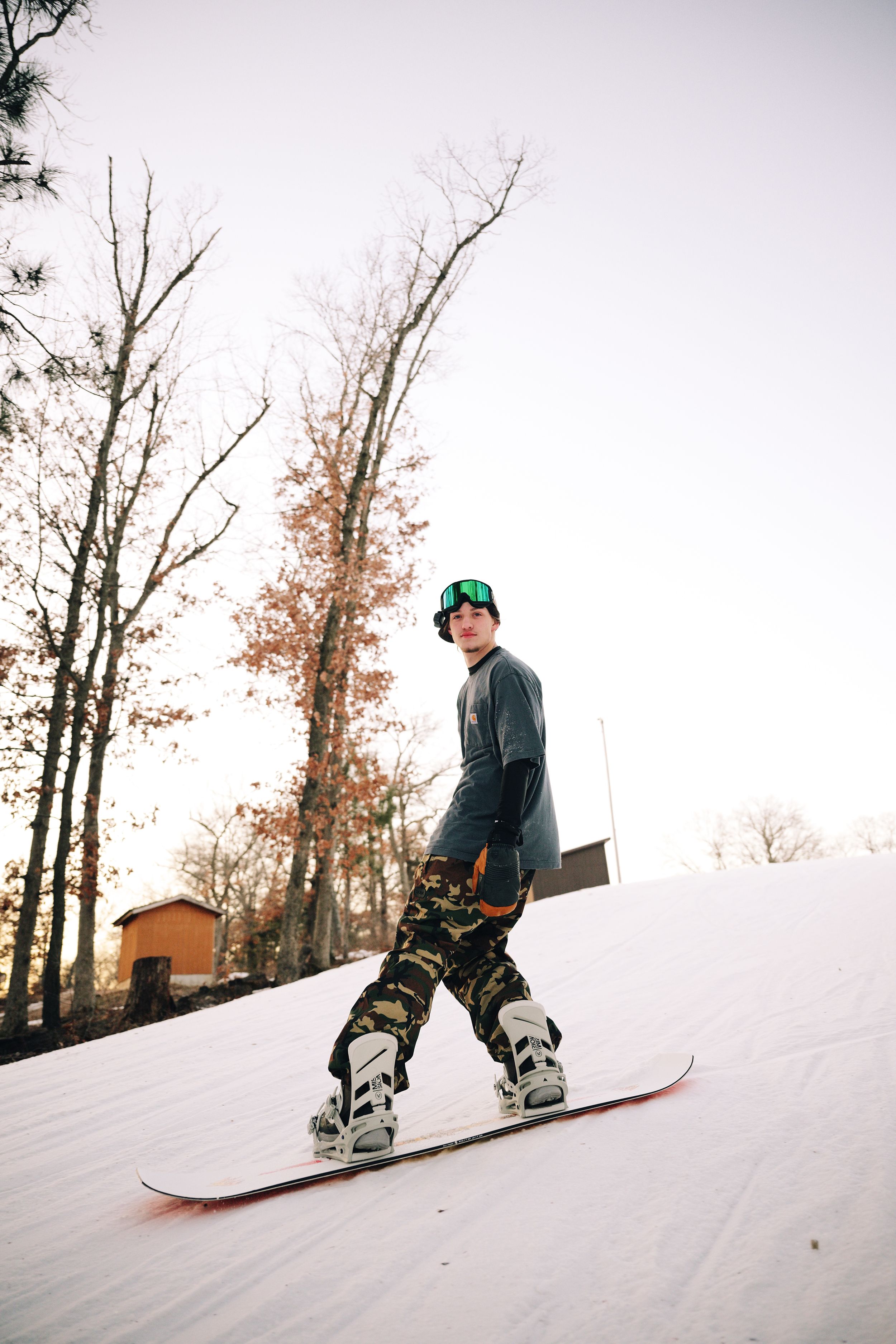 Young snowboarder in camo pants and green goggles riding down a snowy slope among autumn trees.