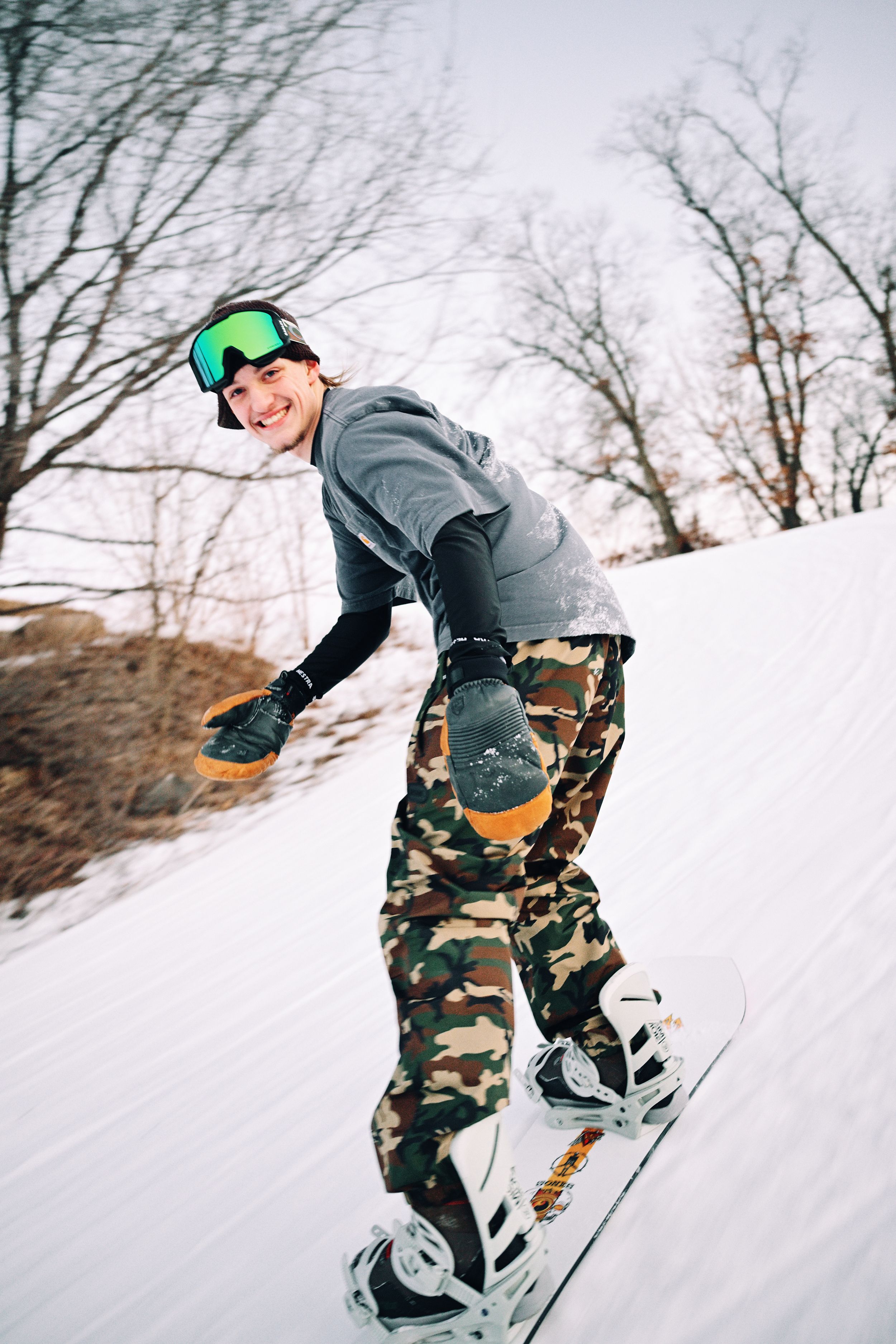 Smiling snowboarder in camo pants and green goggles carving down a groomed snowy ski slope.