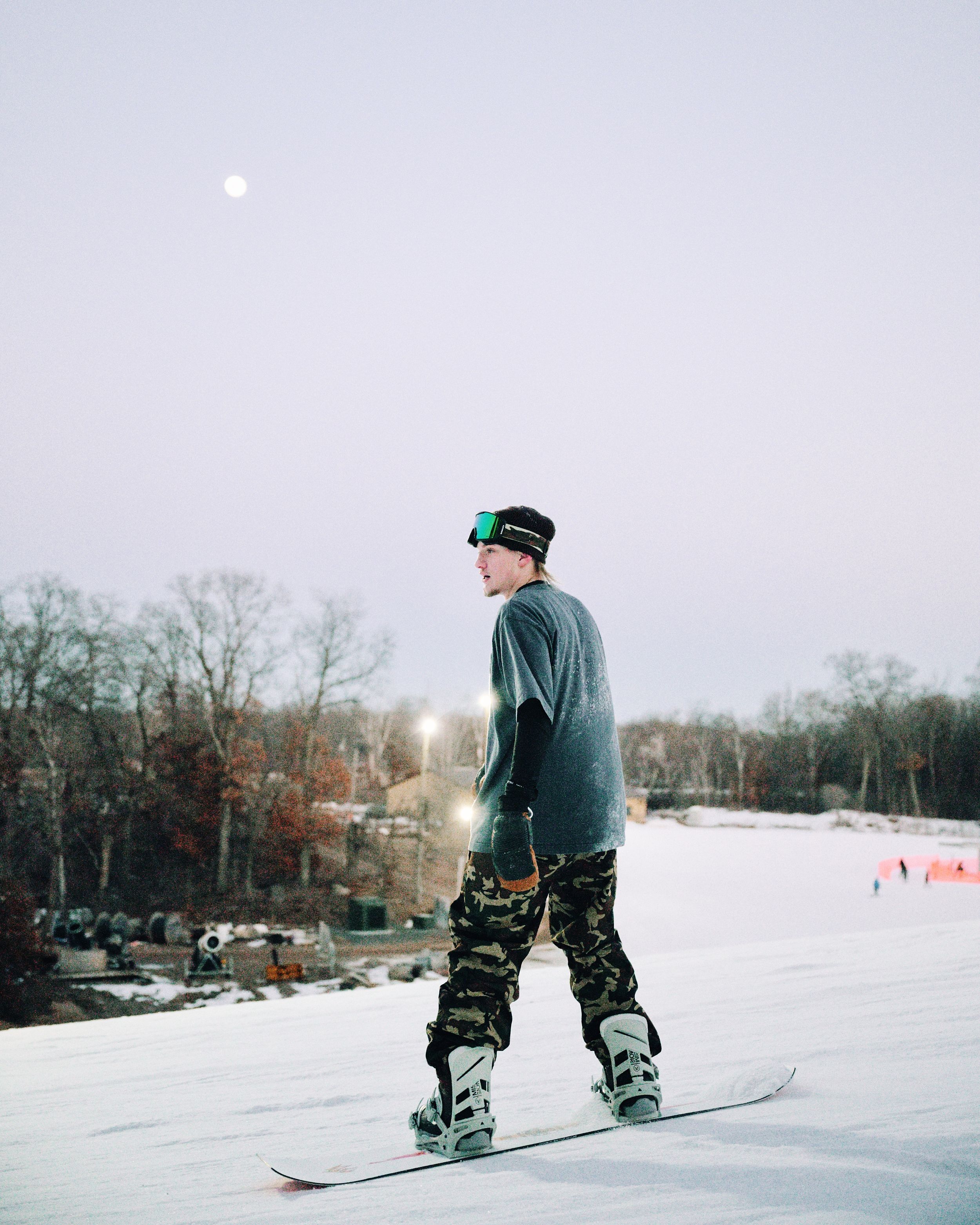 Snowboarder in camo pants and green goggles stands on snowy slope under a full moon at dusk.