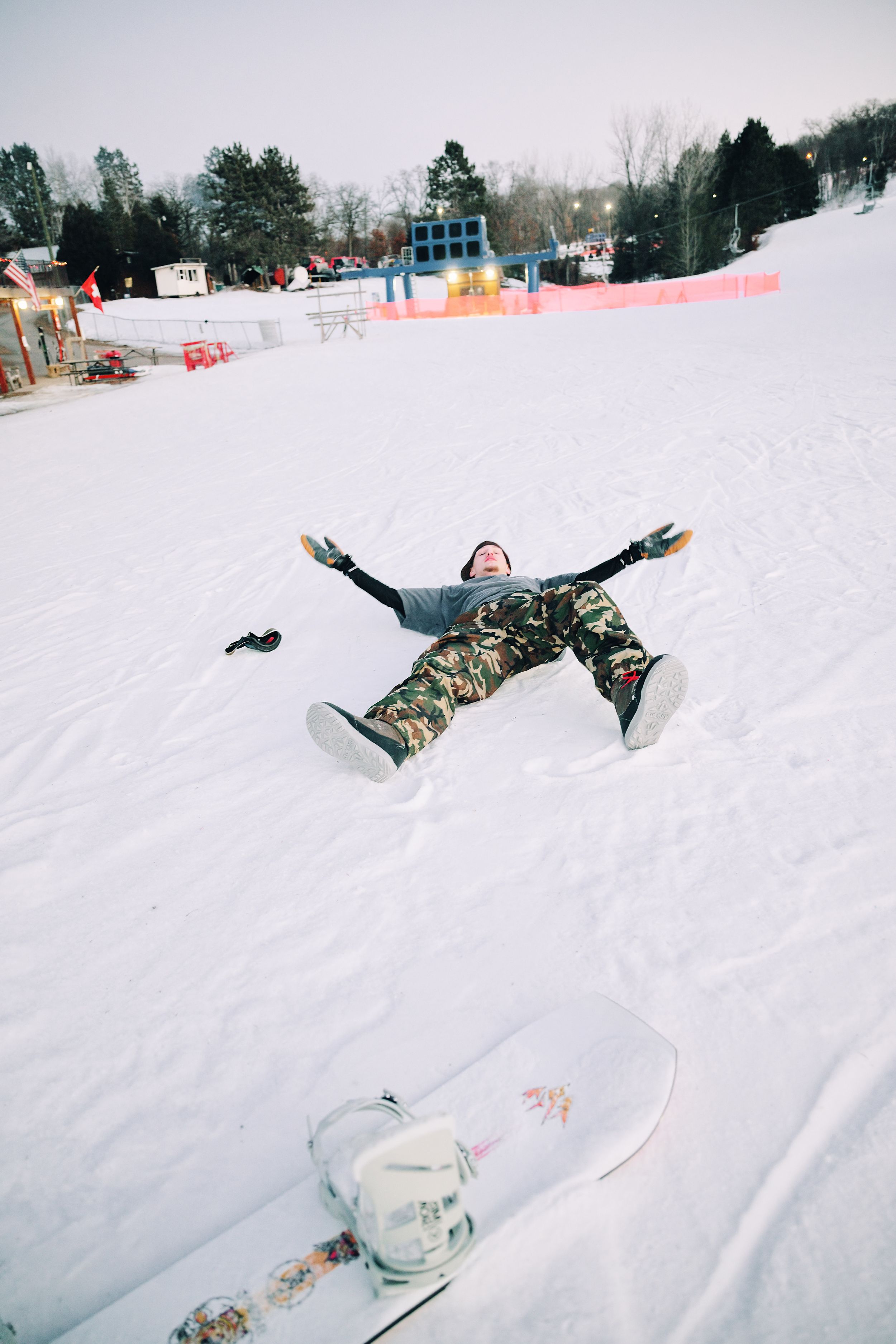 Snowboarder in camo pants lying in the snow making a snow angel with board nearby on ski slope.