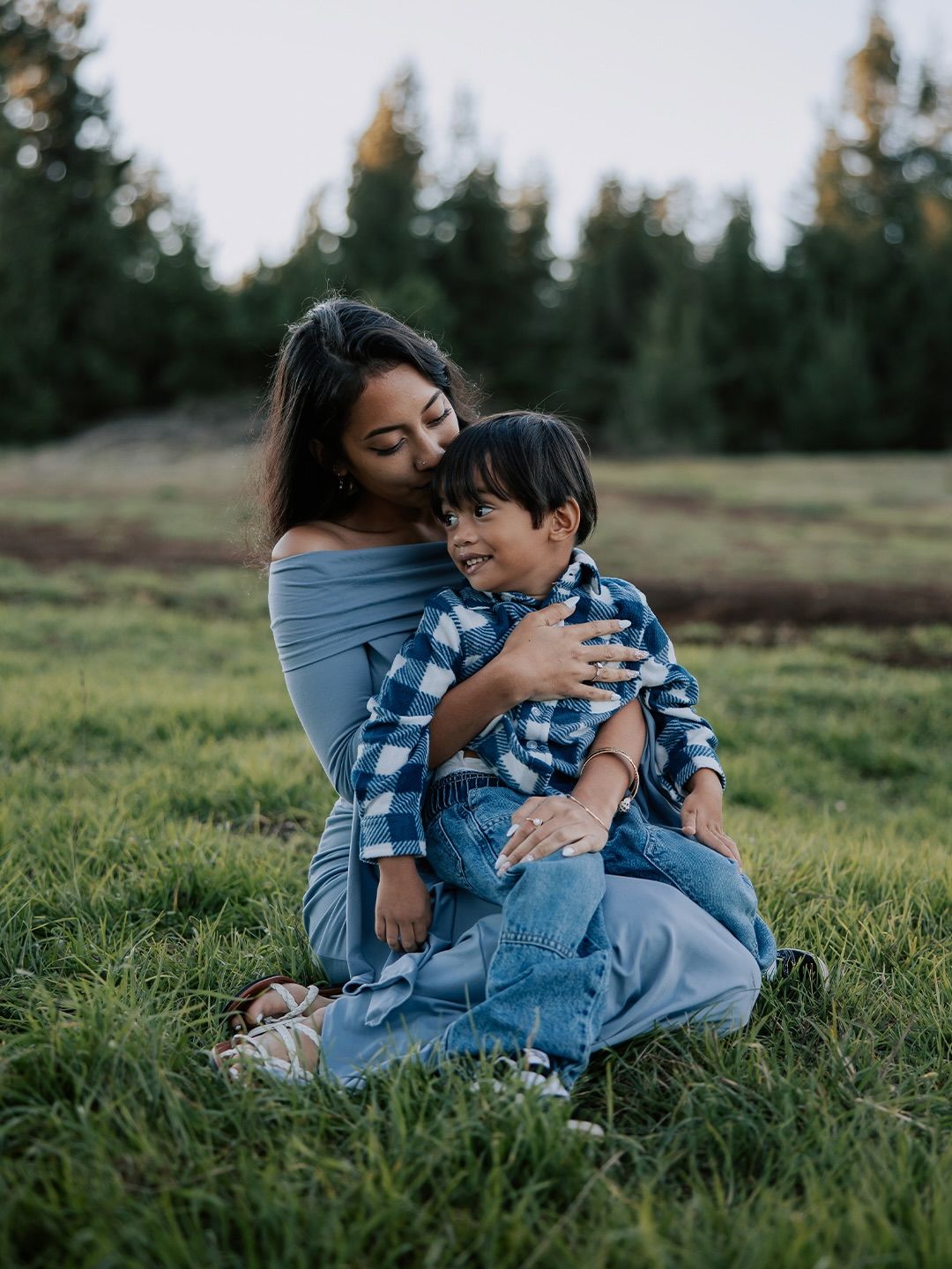 Two people in matching blue plaid shirts share a tender embrace while sitting together in a grassy field at sunset.