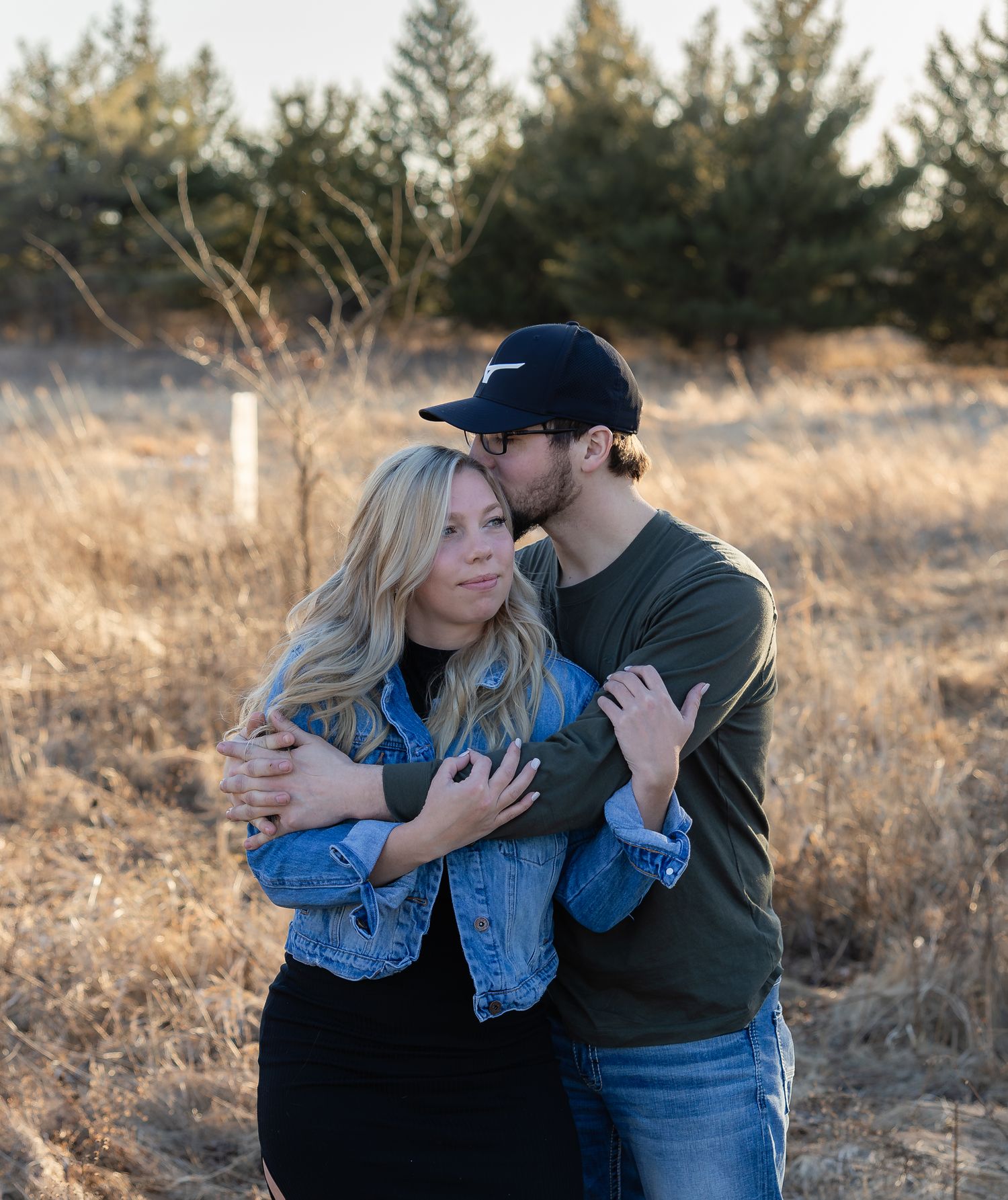 A couple embraces in a romantic outdoor photoshoot against a golden sunset in a rustic field setting.