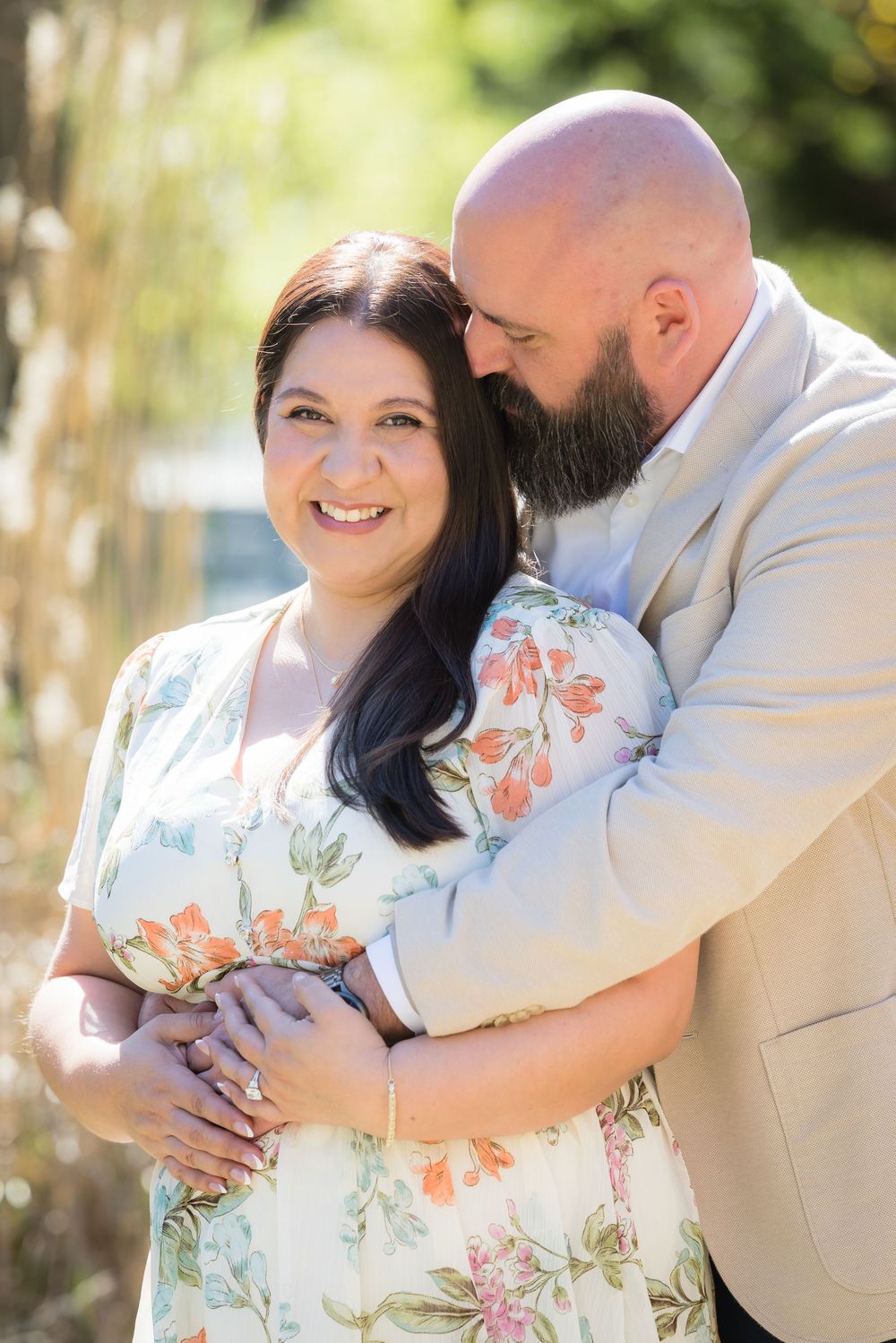 A couple embraces outdoors in romantic maternity photos wearing floral dress and beige suit in natural lighting.