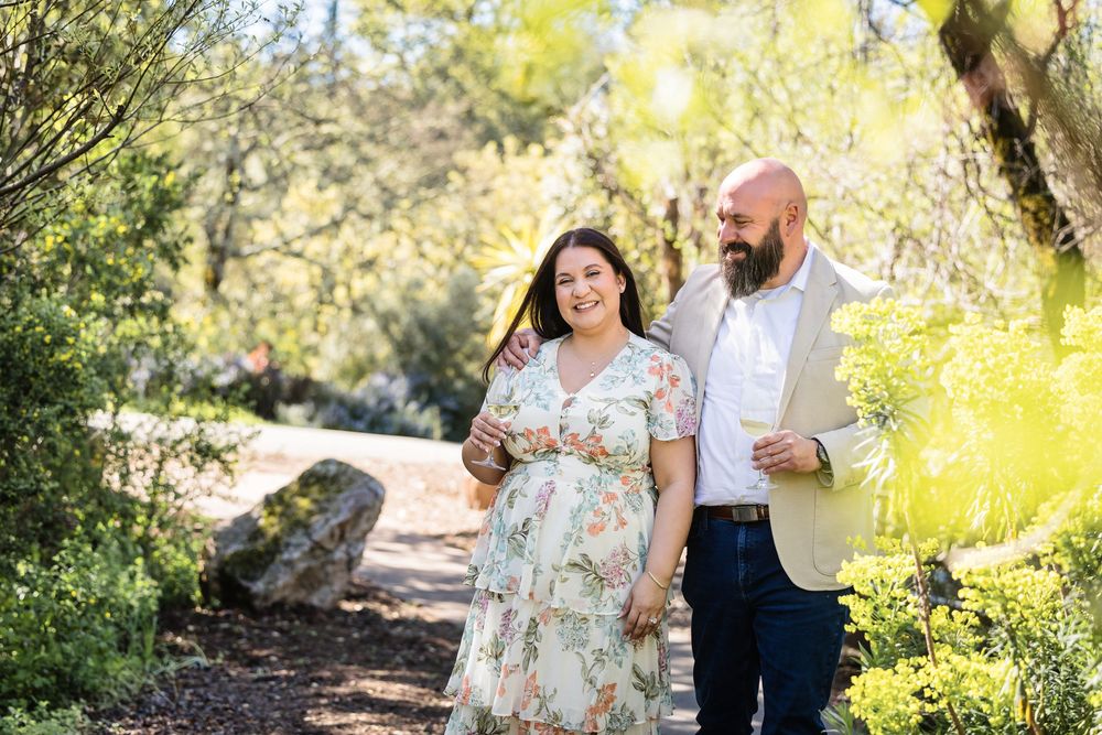 A couple walks together on a sunlit nature trail surrounded by lush greenery and trees.