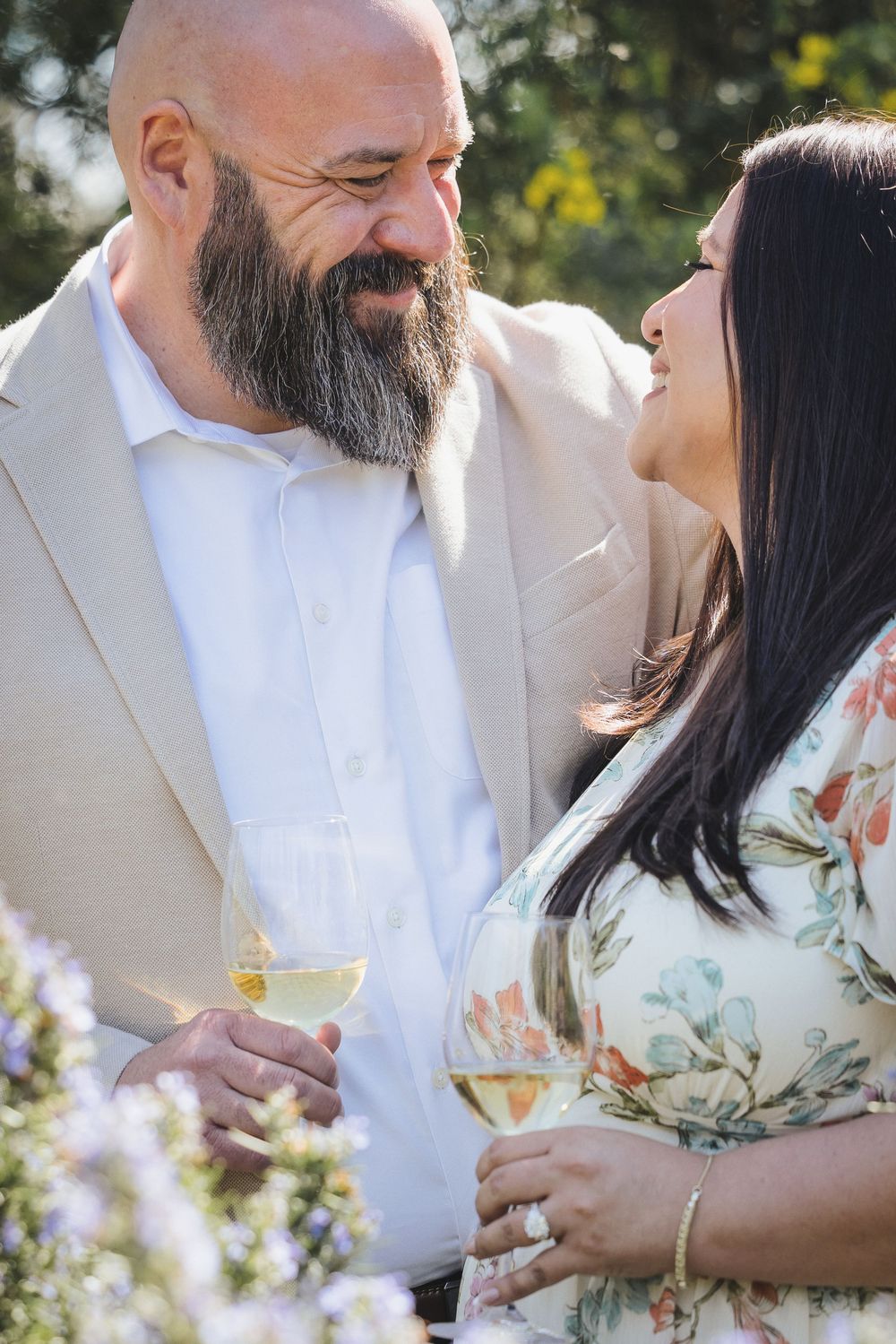 A couple in white and floral attire shares intimate moments while holding champagne glasses in a garden setting.