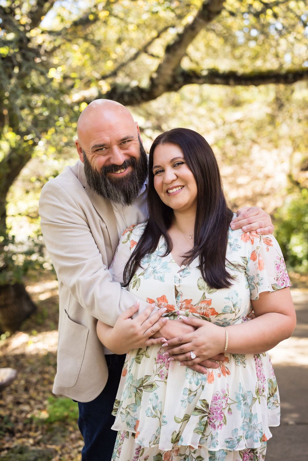 A couple embraces outdoors in a romantic photoshoot among sunlit trees wearing floral dress and beige attire.