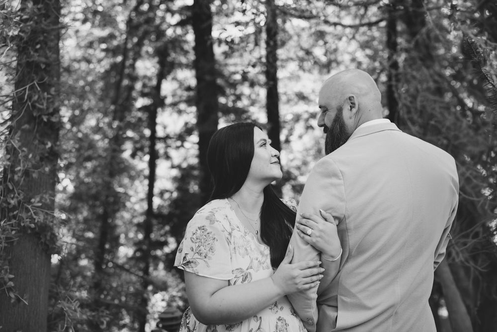 A romantic black and white portrait of a couple embracing in a forest setting among tall trees.