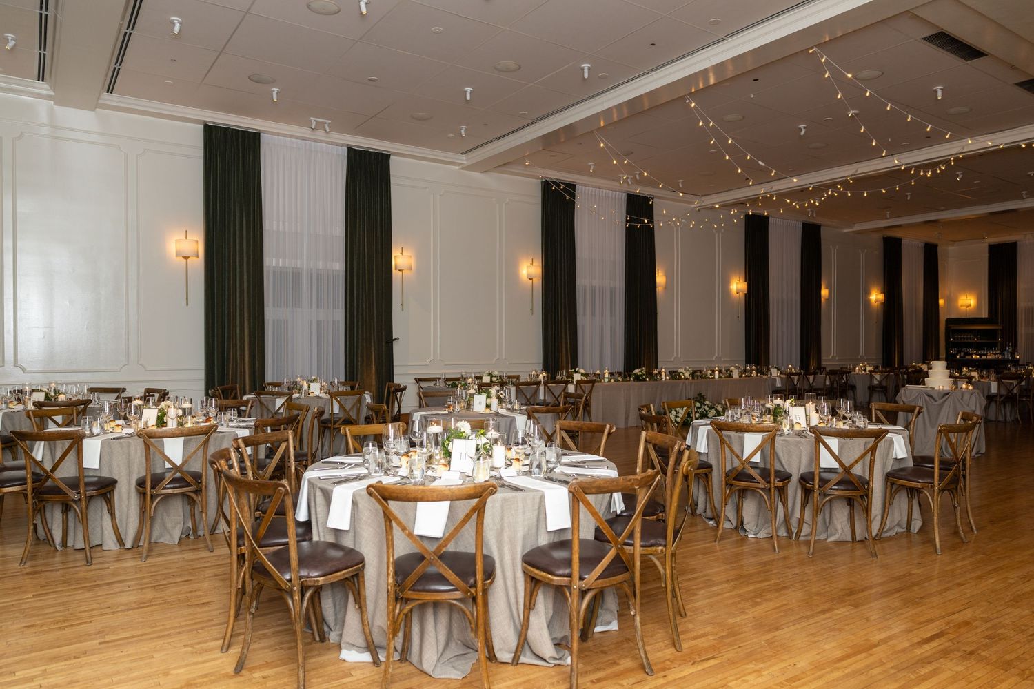 Large banquet hall with white linens and gold chairs set up for an elegant event under twinkling string lights.