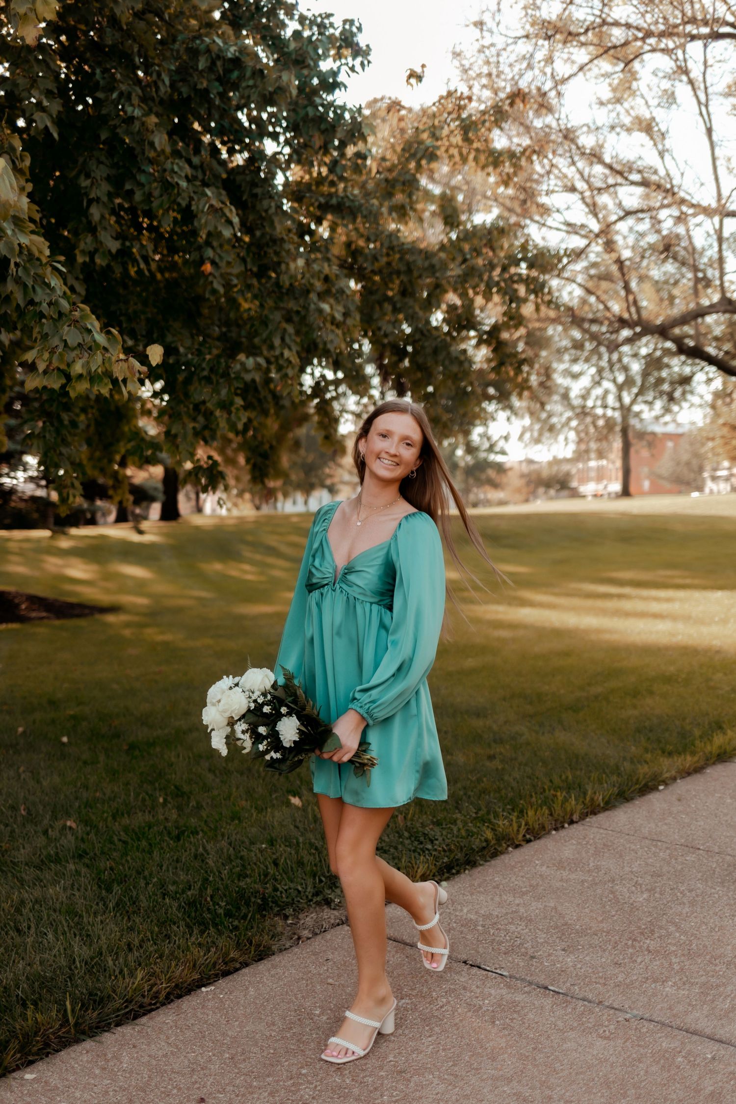 Woman in teal dress holding bouquet while posing on sidewalk near trees.