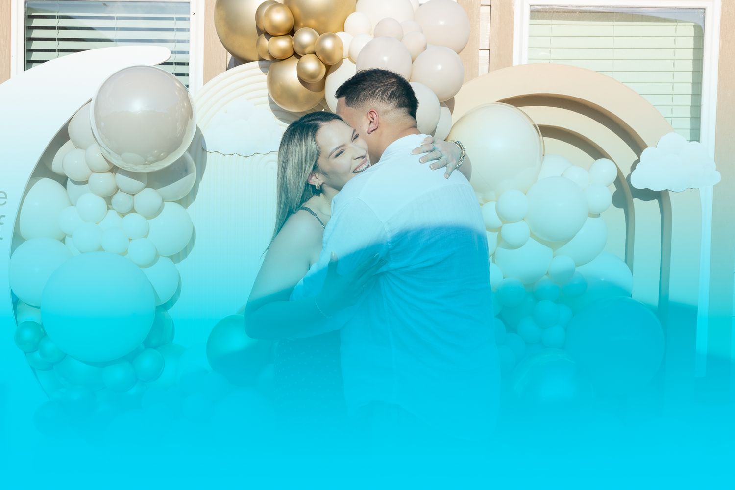 Couple embracing tenderly in front of elegant gold and white balloon arch decoration at a celebration event.