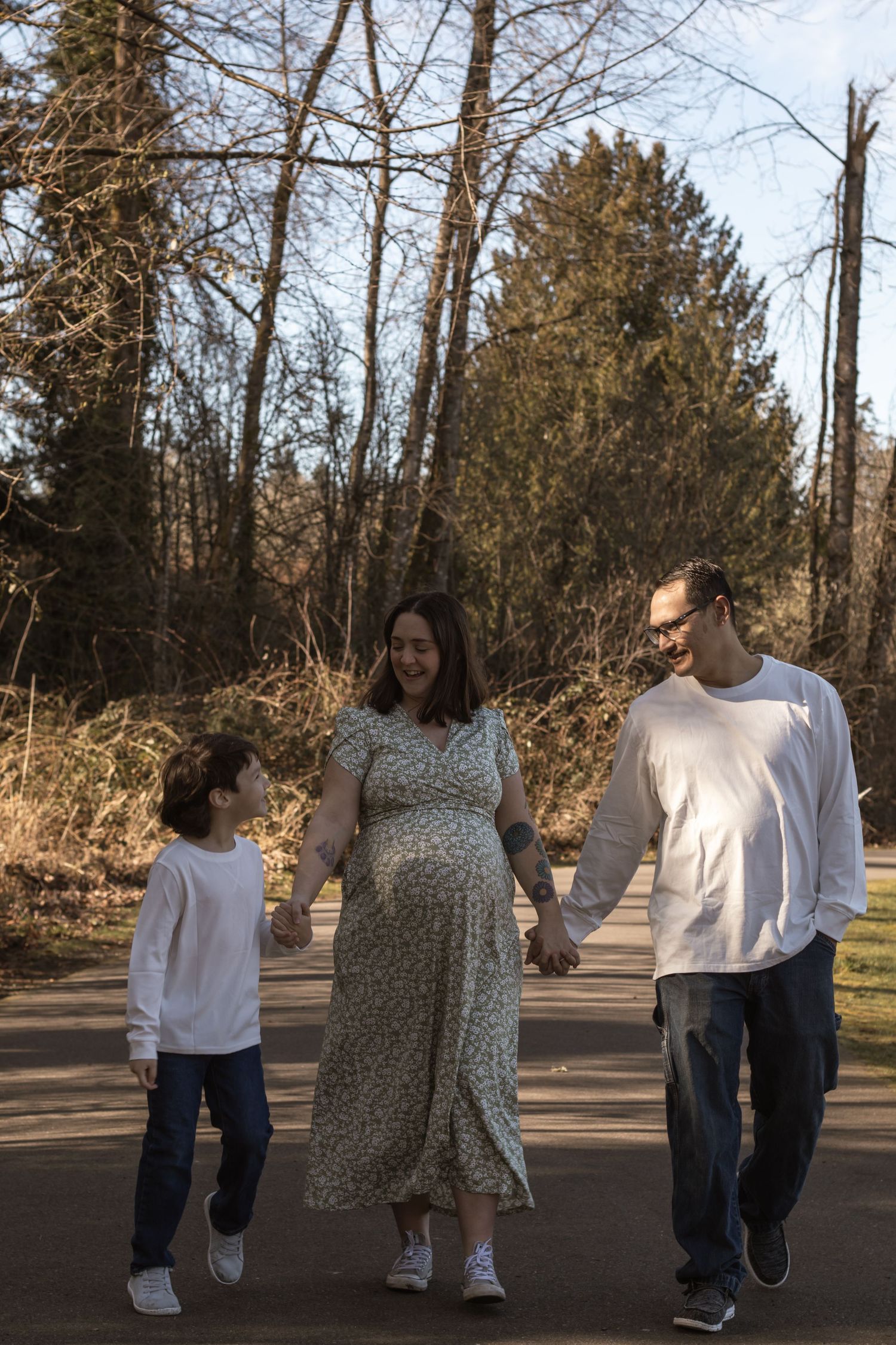 A pregnant woman in a floral dress walks hand-in-hand with her partner and child in a sunny park.
